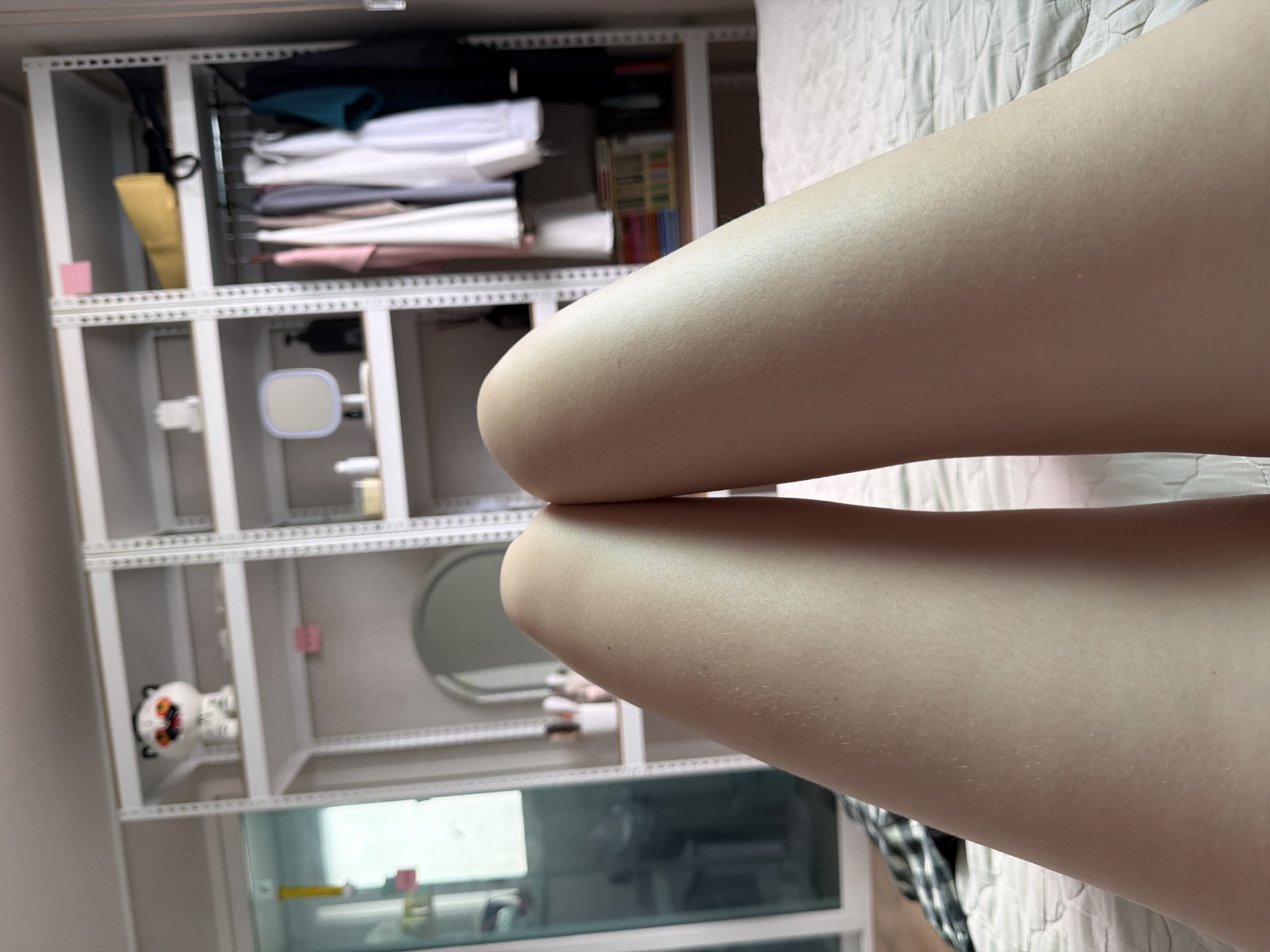 The image shows a close-up view of a person's legs, positioned centrally in the frame and lying on a bed with a quilted white comforter. The legs appear smooth and lightly tanned, with some very fine hairs visible. Behind the legs, a room is visible, featuring a tall, white metal shelving unit. On the shelves, various items are arranged, including clothing hanging on hangers in a closet section, a yellow bag, a small white figurine resembling a tiger, a makeup mirror with lights, and bottles of what appear to be cosmetics or toiletries. To the left of the shelving unit is a glass-fronted cabinet containing some unseen items. The overall impression is that of a bedroom or dressing area in a residential setting. Based on the context provided, this scene is located in Dongducheon-si, South Korea.