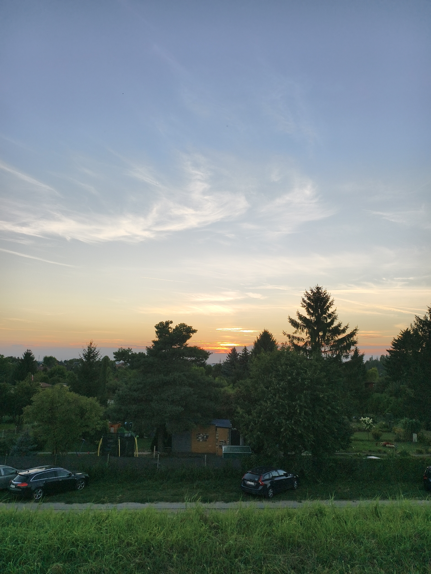 The image depicts a sunset scene from a slightly elevated perspective, overlooking a residential area with lush greenery. The sky is a gradient of soft blues, whites, and oranges, with wispy clouds stretched across it. In the foreground, a strip of vibrant green grass lines the bottom of the frame, suggesting a natural embankment or park.
Beyond the grass, a dark-colored car is parked on a gravelly surface, with another car partially visible to its left. Further back, a collection of trees of varying heights and types dominate the landscape, creating a dense canopy. Among the trees, a small playground structure with yellow and green elements is visible, hinting at a family-friendly environment. A small, rustic-looking building with a yellow facade and a green roof is also present, possibly a garden shed or playhouse. In the distance, more houses and trees are visible, suggesting a suburban or semi-rural setting within the city. The overall impression is one of tranquility and the end of a day.