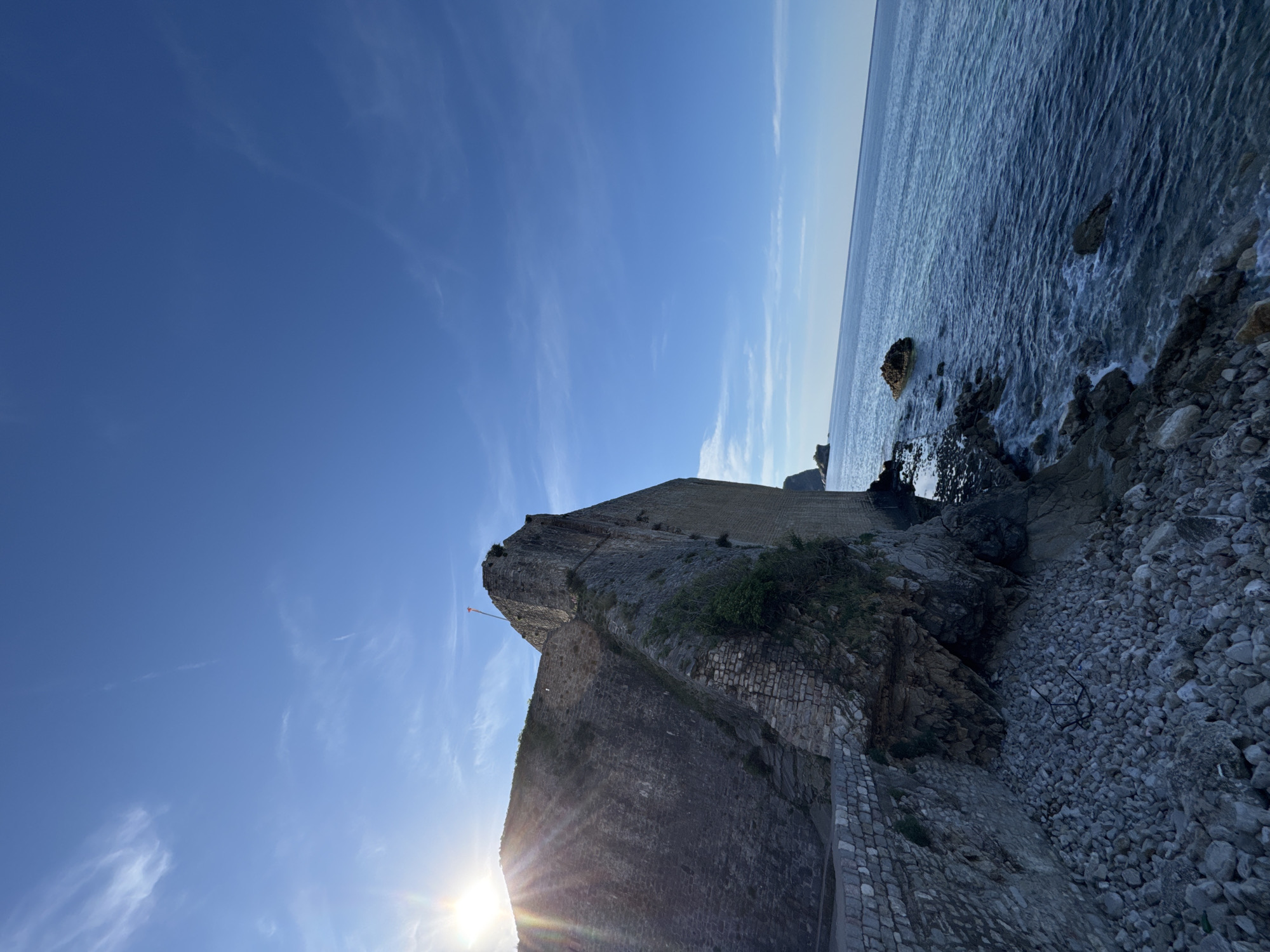 The image shows a stone fortress, likely part of the old city walls of Budva, Montenegro, against a bright blue sky and the Adriatic Sea. The fortress is constructed of grey stone and has multiple levels. The sea is on the right side of the image, with waves crashing against the rocky coastline. The sun is visible in the lower left corner of the image, with sun rays going across the image. A small flag is present on top of the fortress.