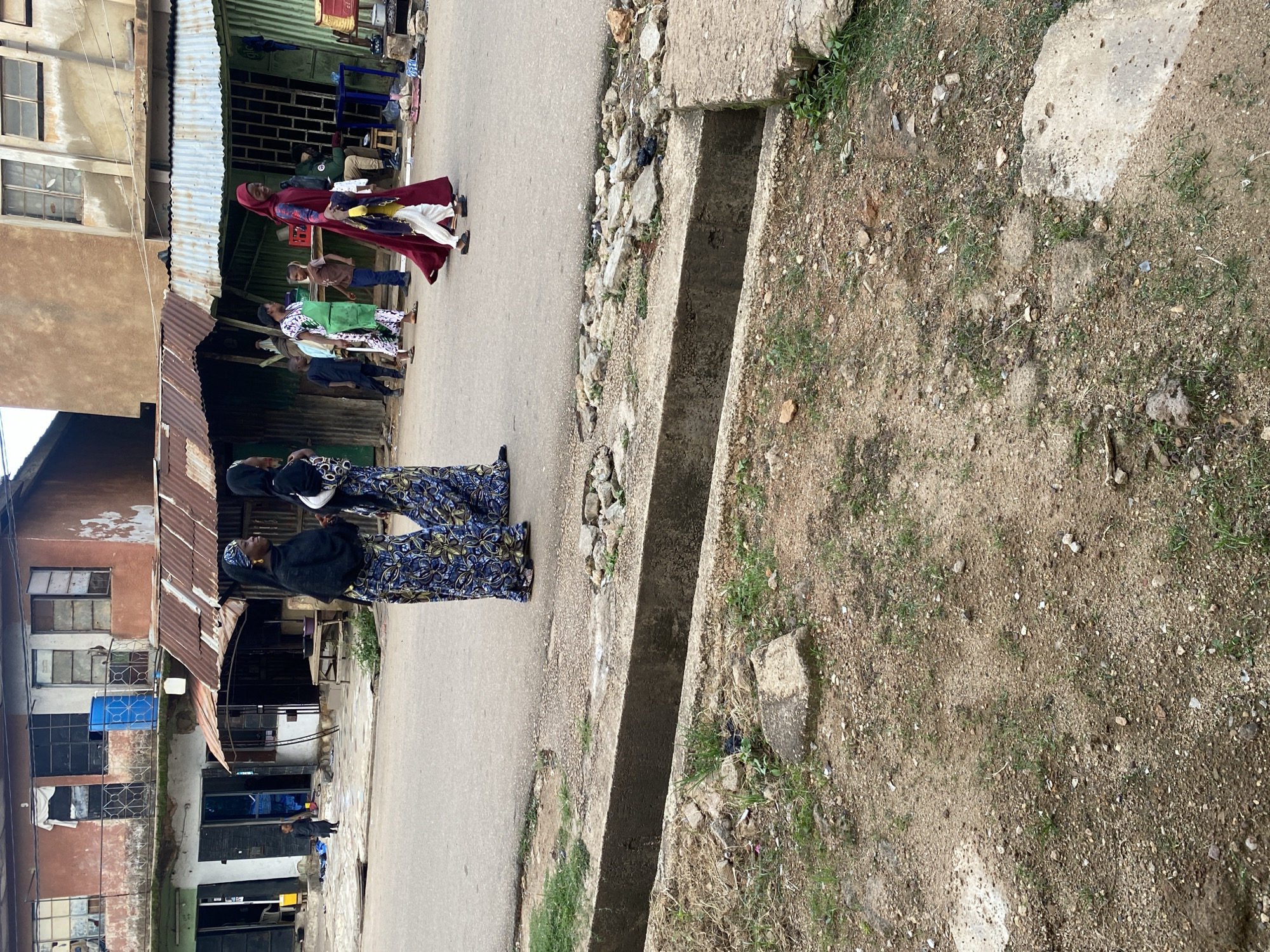 In Jos, Nigeria, on a street, two women in vibrant, patterned attire walk together, their faces turned towards each other in conversation. Behind them, a group of children and other adults move along the uneven pavement. A red and white tent covers a stall with goods, and a man sits at a table under a green awning. The scene depicts a typical street in Jos, with its bustling activity and colorful local culture.