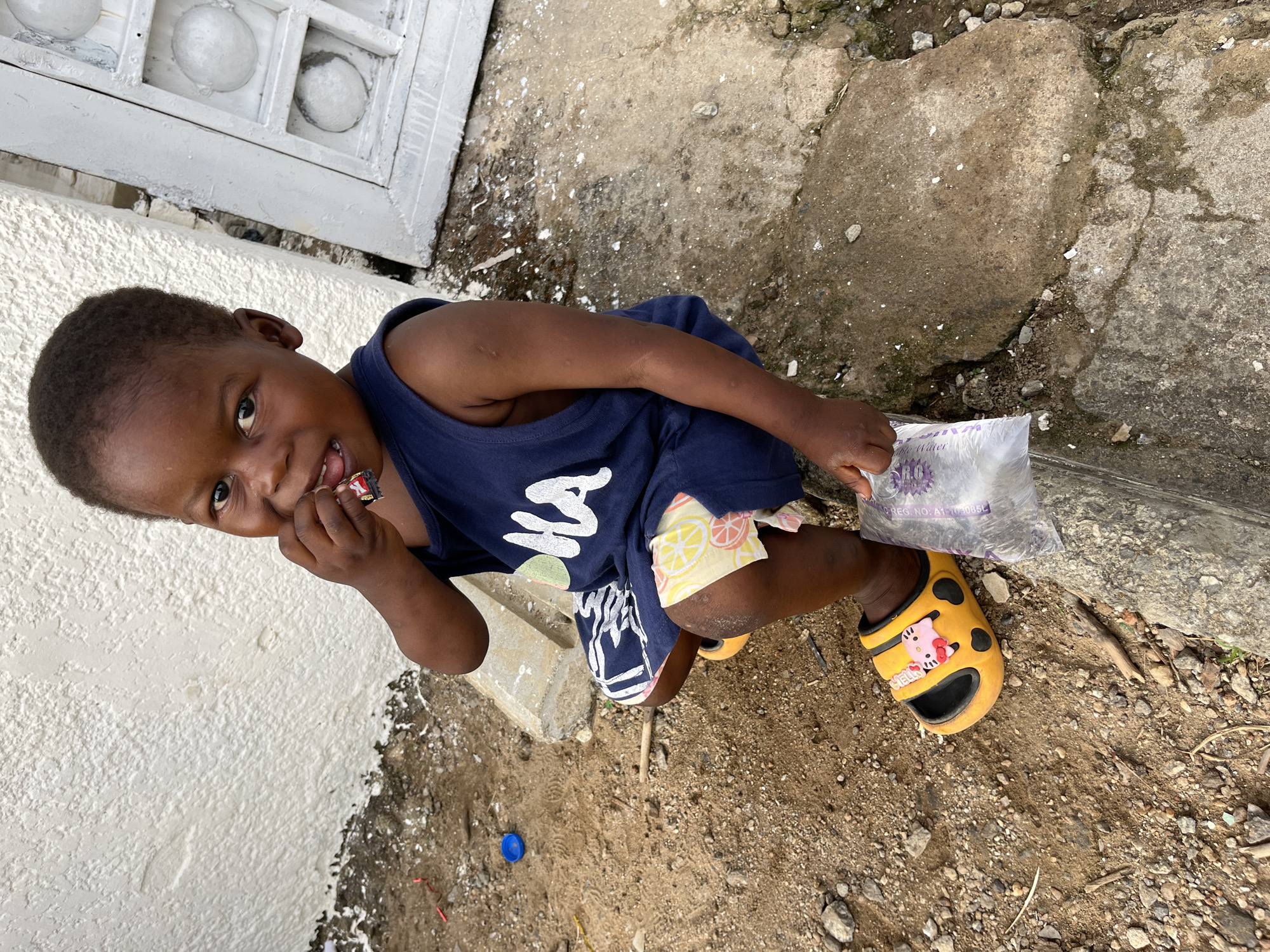 A young child, possibly male, stands in an exterior setting. He wears a dark blue tank top with a white graphic, patterned shorts, and one yellow sandal, possibly Crocs, with a "Hello Kitty" embellishment. In his left hand, he holds a candy bar, while his right hand grasps a plastic bag of water. The environment appears to be a concrete area adjacent to a white wall with a window-like structure. The ground around the child is dirty, with scattered debris, including a blue bottle cap. It is not possible to confirm from the image the location in the city of Abuja, Nigeria.