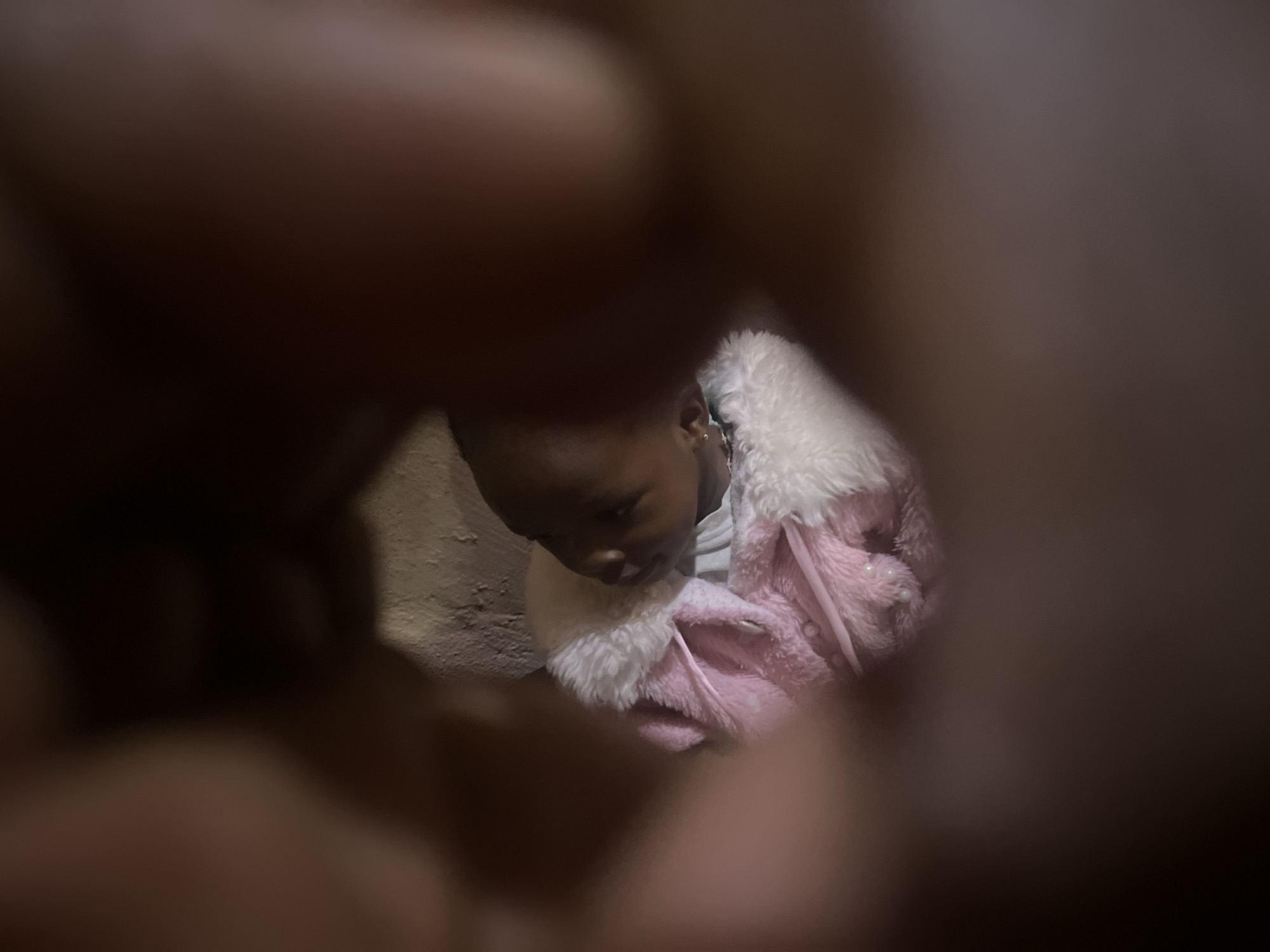 A child wearing a pink and white jacket is partially visible through a circular frame created by blurred fingers. The setting appears to be indoors, with a wall visible behind the child. It is impossible to confirm the location of Gura Top, Nigeria with the given information.