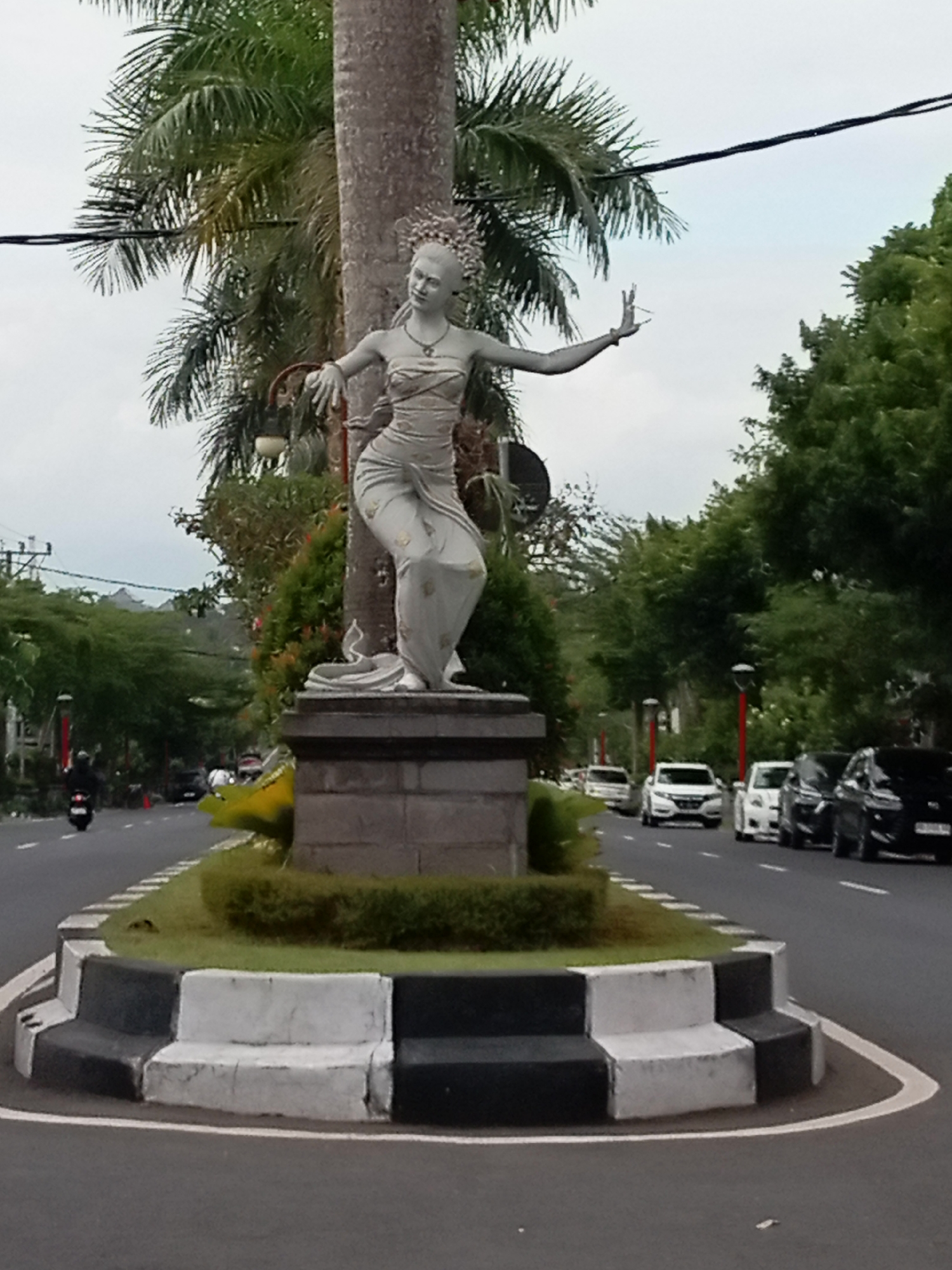 An eye-level, full outdoor shot captures a statue of a woman in a dancer's pose, positioned at a traffic island in an urban area. The statue, which appears to be made of gray stone, is adorned with a floral headdress and a patterned dress. It stands on a rectangular stone pedestal, surrounded by green bushes. The traffic island itself is bordered by a curb painted with alternating black and white blocks, and a white line indicates the edge of the road.
The road curves around the island, with several cars and a motorcycle visible. In the background, lush green trees and palm trees line the street, their foliage creating a vibrant contrast with the overcast sky. A power line stretches across the upper portion of the image. The overall impression is one of a roadside monument in a tropical or subtropical environment, where traffic flows around a cultural or artistic landmark.