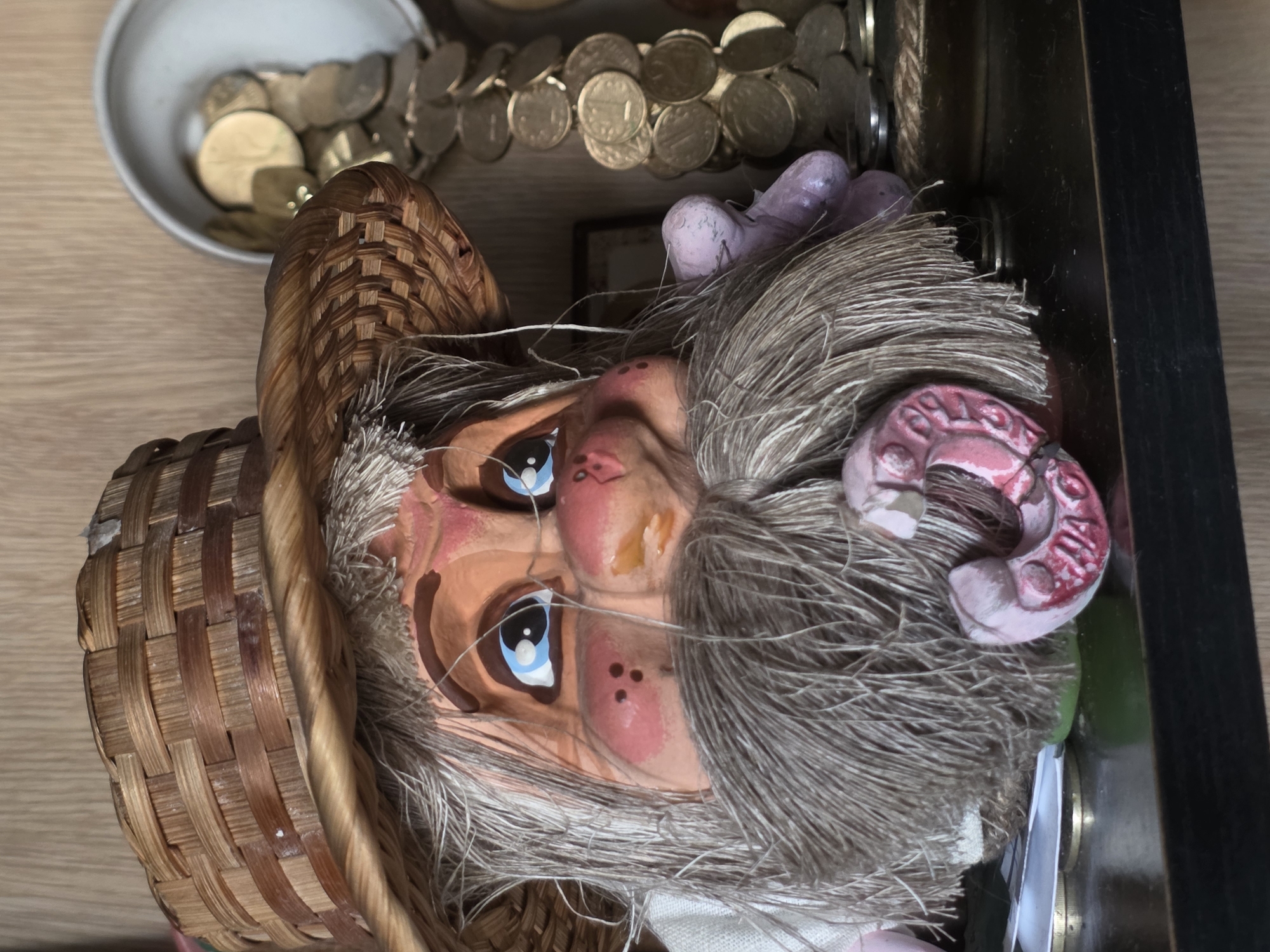 The image shows a close-up, rotated view of a doll's face. The doll has a painted face with blue eyes, rosy cheeks, and a grey, wispy beard. It is wearing a woven straw hat. The doll appears to be an older, possibly gnome-like character. Behind the doll, there are several objects visible, including a bowl filled with coins and a string of coins arranged in a curve. The background includes a wooden surface and a dark wooden frame. The lighting suggests it is daytime, and there are no indications of specific weather conditions. The scene is likely a still-life arrangement, possibly a collection of decorative items. No visible text is present in the image.