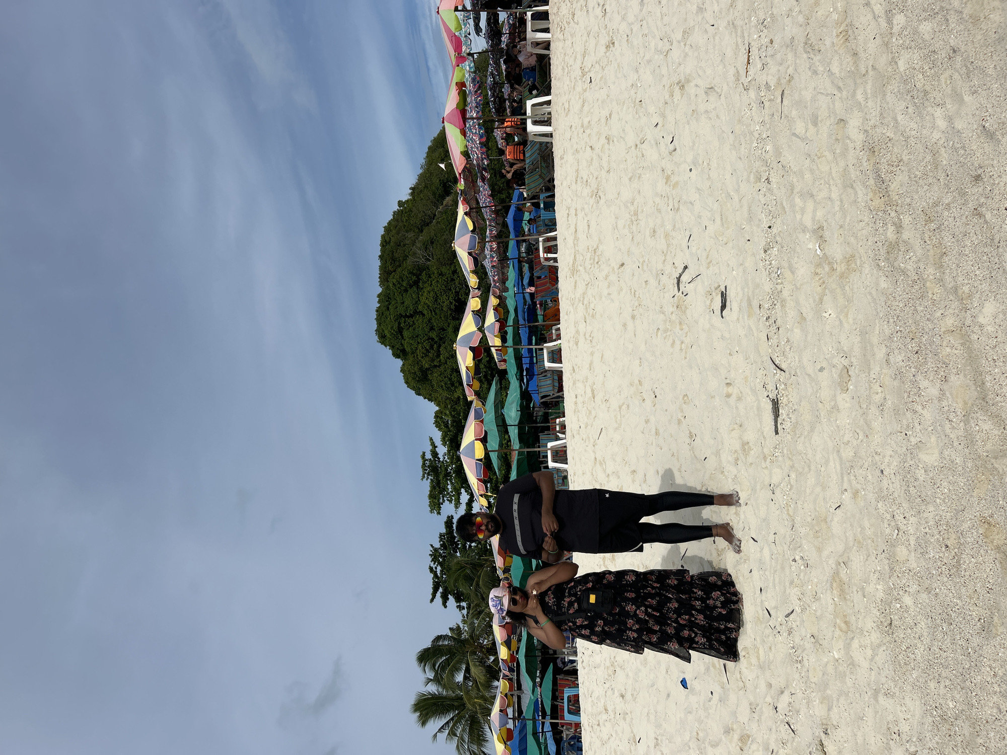 This image captures a couple enjoying a day at the beach in Tambon Phru Nai, Thailand. The scene is dominated by the sandy shore, with a line of colorful beach umbrellas and chairs set up in the background. Lush green trees and a hillside form the backdrop, indicating a tropical or subtropical location. The sky above is partly cloudy, suggesting it's daytime with perhaps some diffused sunlight.
The couple is positioned on the sand, with the man standing and the woman in front of him. The man is wearing a black t-shirt and black leggings, while the woman is in a floral print dress. Both appear to be wearing sunglasses, shielding their eyes from the sun. They seem relaxed and engaged with each other, possibly posing for a photo or simply enjoying each other's company.
The beach appears to be a popular spot, with numerous umbrellas and seating visible. The presence of palm trees hints at the tropical climate. The overall mood of the image is one of leisure and vacation, characteristic of a beach setting. There is no visible text in the image.