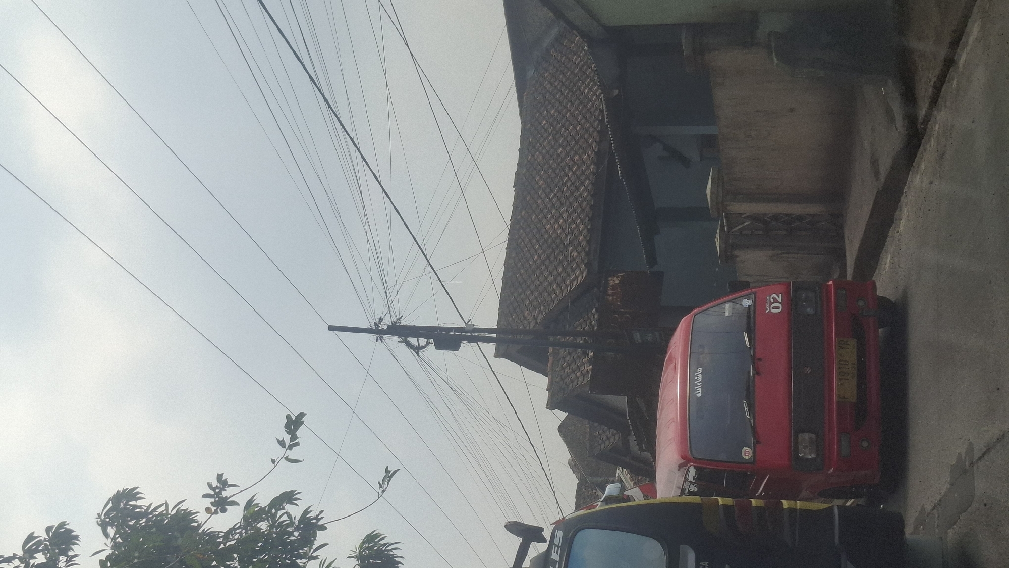 In Sirnagalih, Indonesia, under an overcast sky, a street scene unfolds with a red Suzuki Carry van prominently featured in the foreground. The van, with license plate "F 1910 YR", is parked on the right side of a paved road, facing forward. Its windshield displays Arabic calligraphy. To the left of the van, the rear portion of a yellow and black vehicle is visible. Behind the vehicles, a row of houses with tiled roofs and weathered walls lines the street. A utility pole with a complex network of overhead wires stands tall amidst the residential structures. Trees with green foliage are present on the left side of the frame, reaching towards the sky. The overall environment suggests a residential area within a city.
