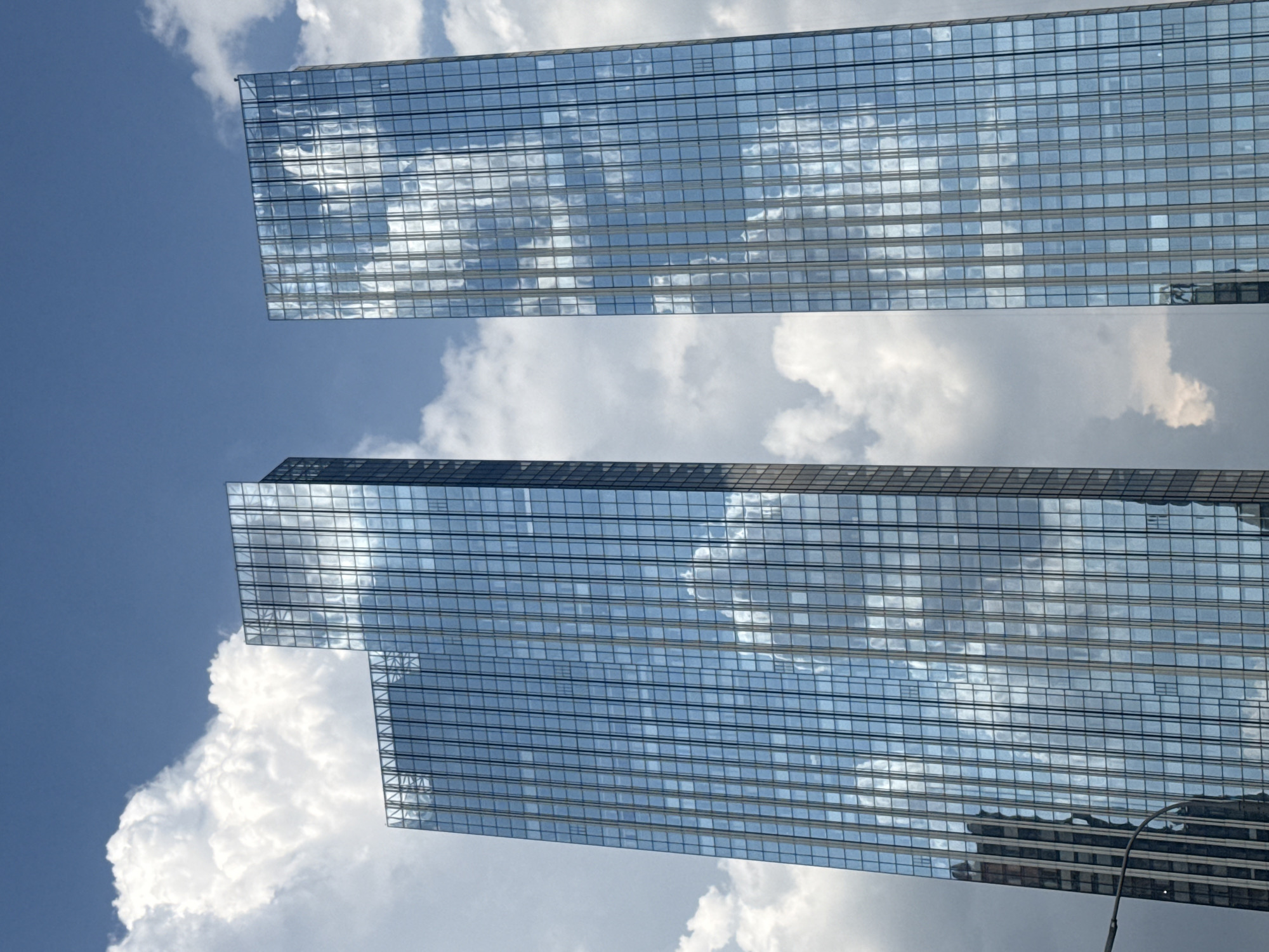 The image shows two modern glass skyscrapers against a cloudy blue sky. The buildings are tall and rectangular with a grid-like pattern of windows that reflect the sky and clouds. The skyscraper on the left is slightly shorter and appears to be set back from the one on the right. A street lamp is visible in the lower left corner, partially obscuring the base of the left skyscraper. The overall scene is urban and contemporary.