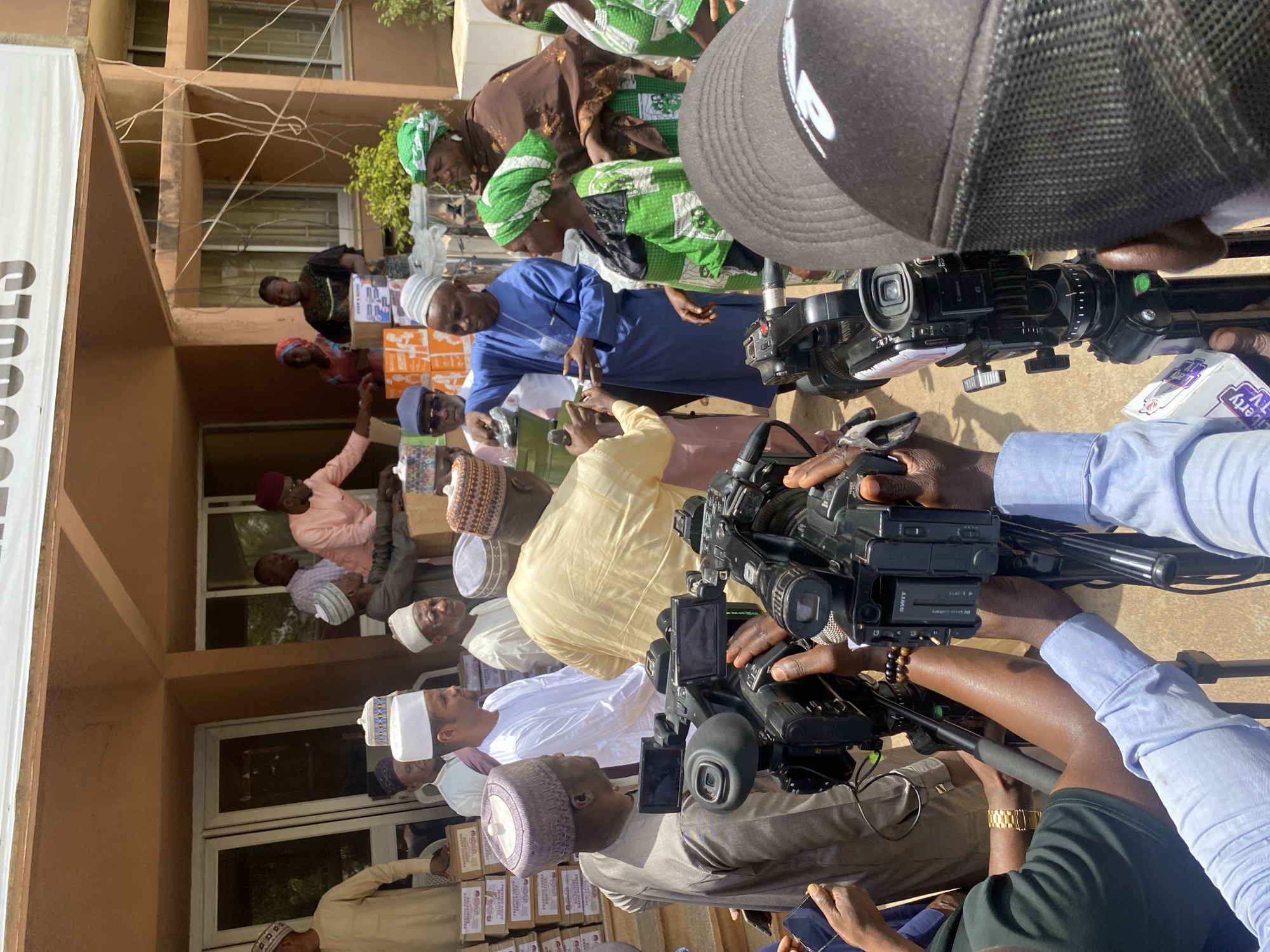 The image captures a scene in Unguwar Rimi, Nigeria, where a group of people, predominantly men, are gathered outdoors in front of a building. Many are wearing traditional Muslim attire, including kufi caps and flowing robes. Boxes are visible, suggesting some sort of distribution or donation event. Several professional video cameras, held by camera operators, are pointed towards the group, indicating media coverage of the event. The environment is a sunlit outdoor area, with the building's architecture visible in the background.