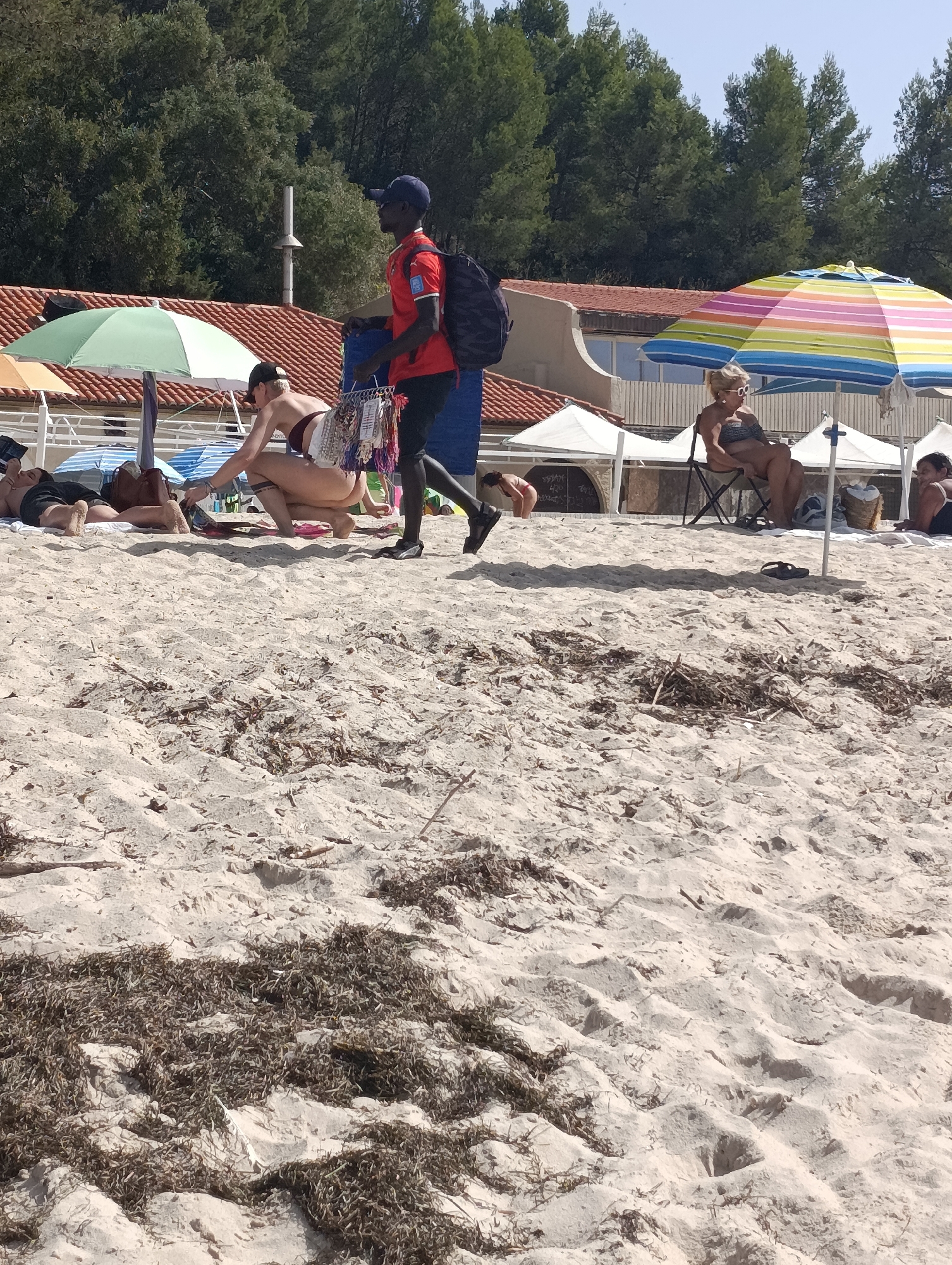 A man in a red shirt and blue cap walks across a sandy beach, carrying a backpack and a basket of goods. He is in the foreground, walking away from the viewer. In the background, several people are relaxing on the beach under colorful umbrellas. The beach is lined with trees and buildings. The scene appears to be a typical summer day at the beach. There is no indication that this scene is in Setúbal, Portugal.