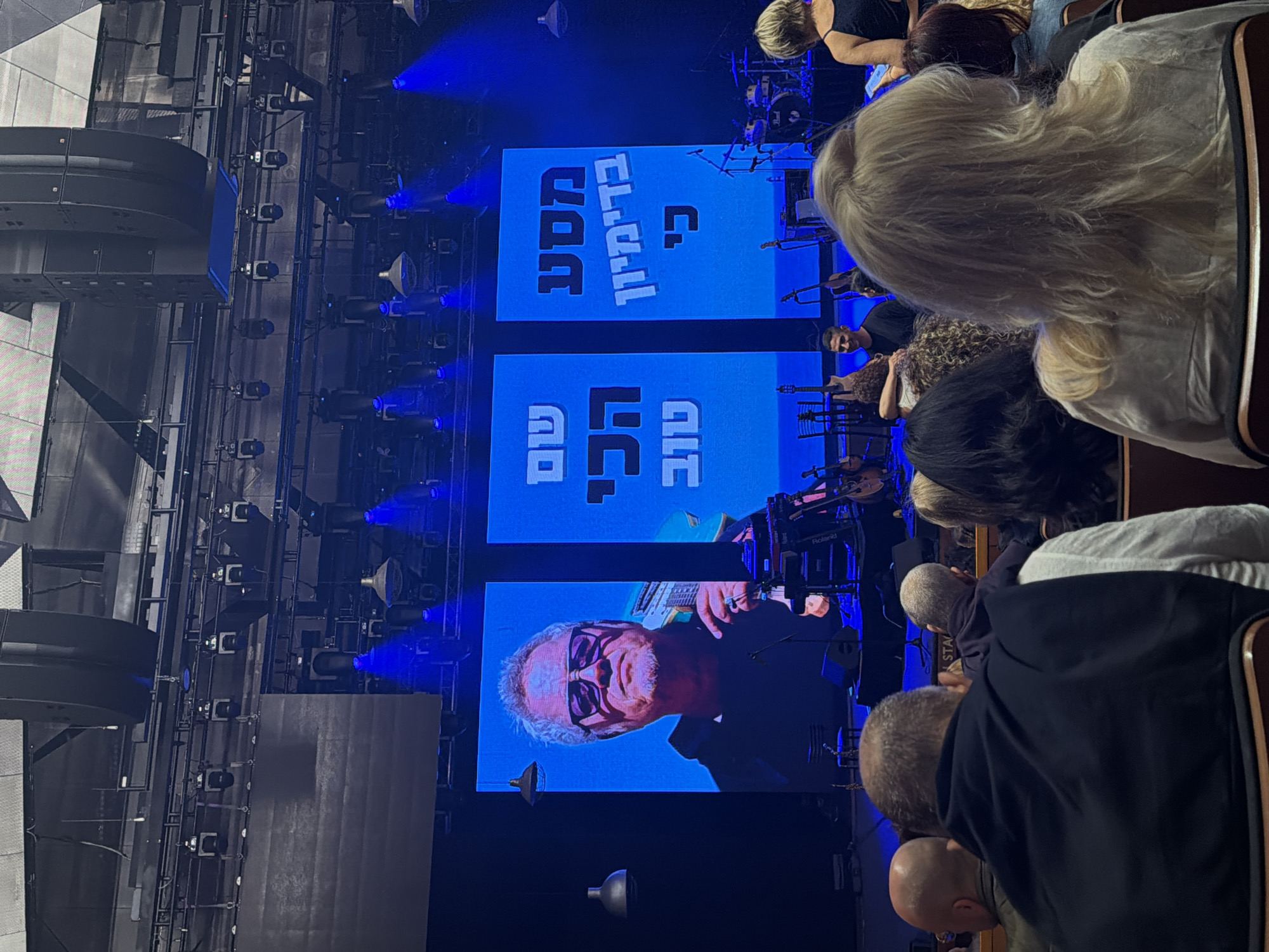 The image shows a concert performance, likely in Tel Aviv, Israel. On the stage, displayed on a screen, is an older man with white hair and glasses. Text is also visible on the screen, written in Hebrew. In front of the stage, an audience is seated, with the backs of several people clearly visible. There is blue stage lighting. Stage rigging and equipment are visible on the left side.