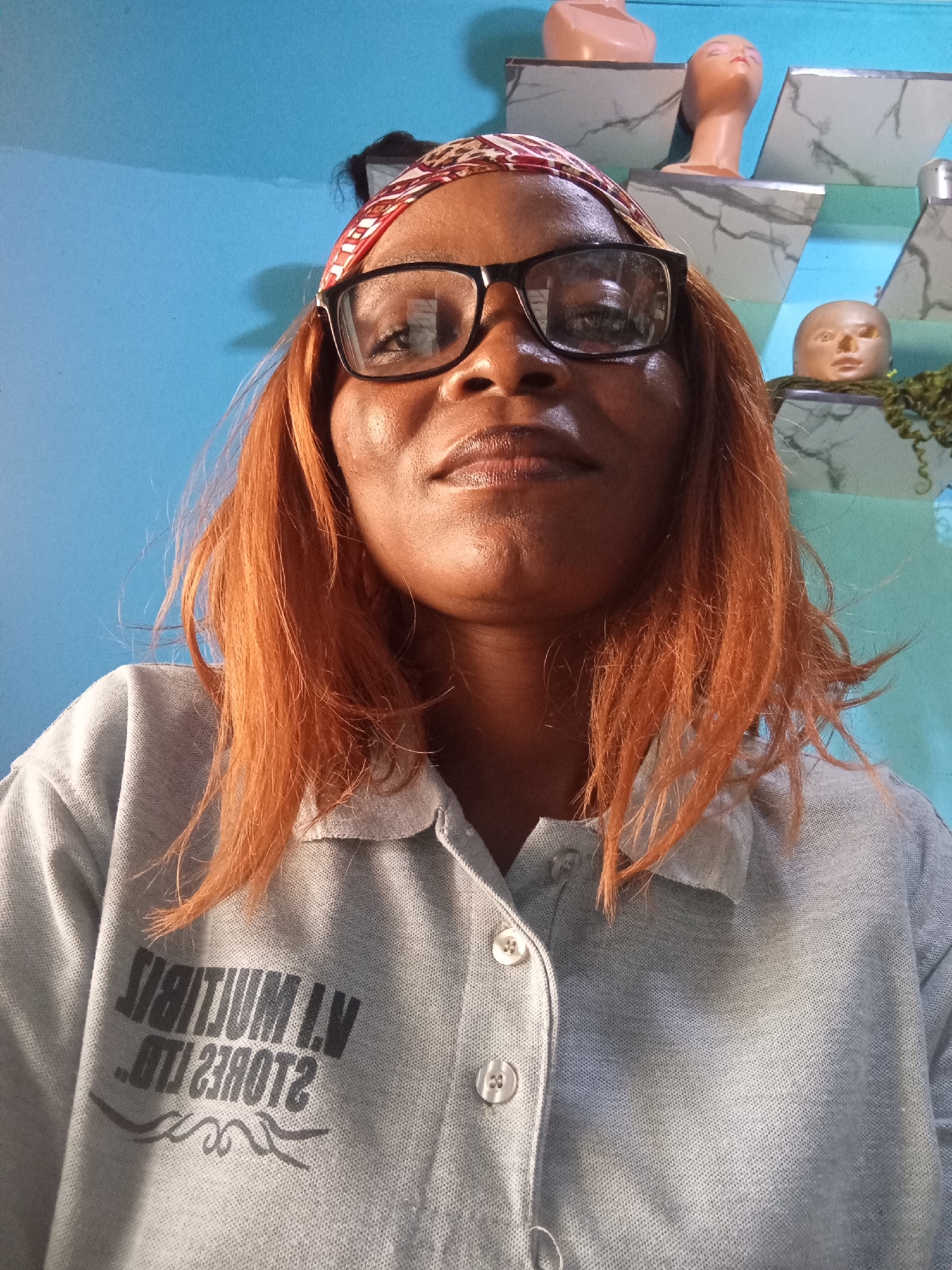 A selfie, taken at a slightly downward angle, features a Black woman with copper-colored hair, a red and white patterned headscarf, and black-rimmed glasses. She is wearing a light gray polo shirt with buttons and a printed logo. The background is a bright blue wall, with shelves holding what appear to be mannequin heads and decorative objects. The overall impression is of an indoor setting, likely a home or shop in Jalingo, Nigeria, given the mention in the prompt. The lighting is bright, suggesting it might be daytime, with sunlight possibly contributing to the highlights on her skin and hair.