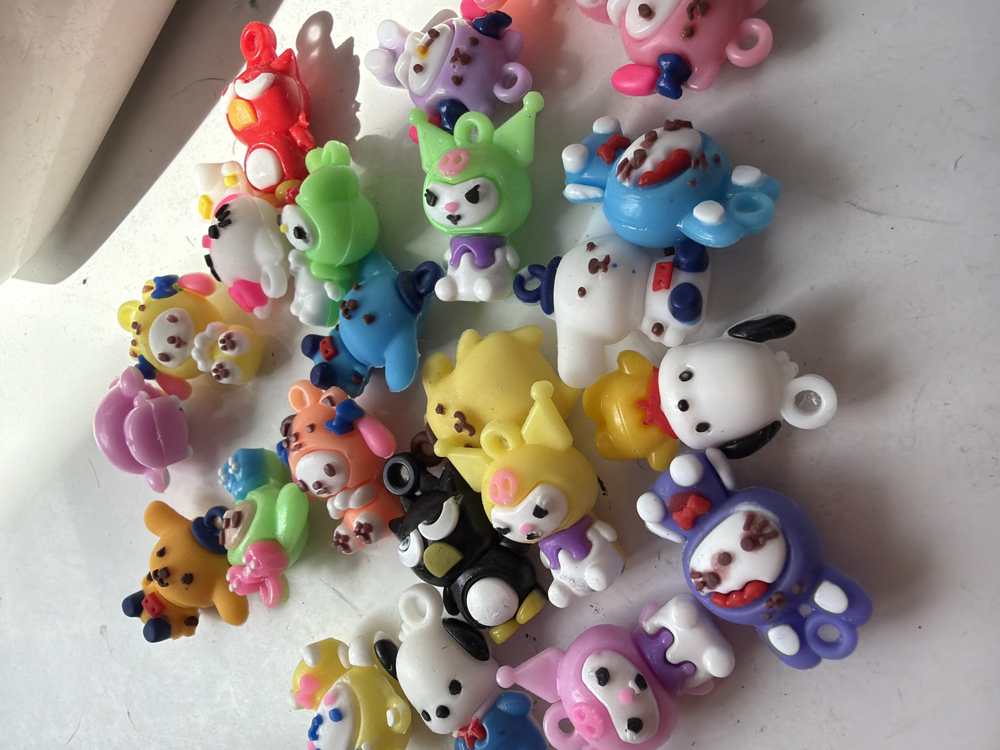 The image shows a collection of small, colorful, cartoon-like plastic charms or figurines scattered on a white surface. The figurines appear to be of various characters or animals, with details such as ears, eyes, and decorative elements. Some are red, green, pink, yellow, black, blue, white, orange and purple. The charms are likely meant for craft projects or decorations. The background is a plain, white surface. There are no indications that this image was taken in the city of Saint Petersburg, Russia.