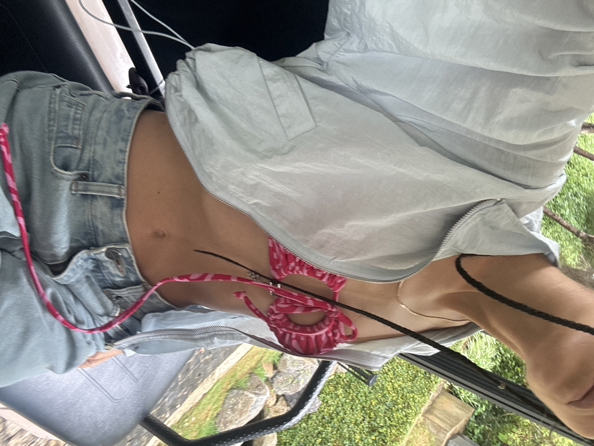An overhead, vertically oriented shot captures the mid-section of a person, likely outdoors in Púng Luông, Vietnam, during the daytime. The individual is wearing light blue, faded denim shorts, a pink and white patterned bikini top with tie details, and a light blue, lightweight, unzipped windbreaker jacket. A thin gold chain necklace is visible around the neck. A pink patterned strap, possibly a lanyard or bag strap, rests across the denim shorts. A black braided cord is also visible on the right side of the frame.
The person's bare midriff is visible between the jacket and shorts. In the background, green foliage, including grass and bushes, and several rocks suggest a natural or semi-natural outdoor environment. Parts of a dark structure, possibly a chair or vehicle interior, are visible in the foreground and along the left edge. The bright natural lighting indicates it is daytime. There is no visible text in the image.
