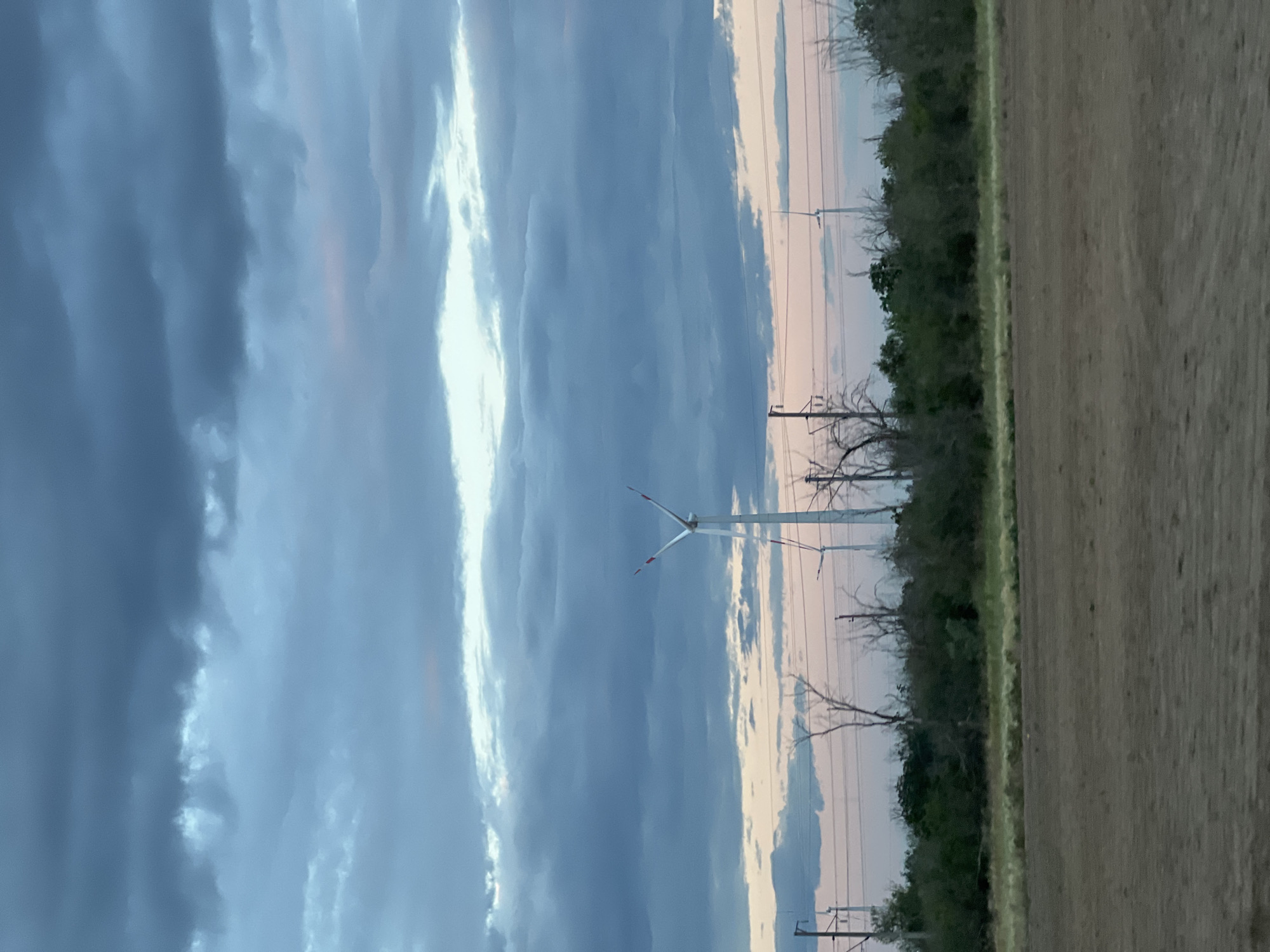 The image depicts a landscape scene with a cloudy sky and wind turbines. The sky is filled with dramatic, dark blue and grey clouds, with a bright white streak of light cutting through them. In the distance, several wind turbines stand tall, silhouetted against the lighter parts of the sky. The turbines have white blades with red tips, and one is prominently visible in the center of the frame.
The foreground on the right side of the image shows a section of a field, appearing to be agricultural land, with a line of trees and vegetation separating it from the sky. The trees are bare of leaves, suggesting it might be autumn or winter. There are also power lines and poles visible, indicating human infrastructure in the area.
The overall impression is one of a vast, open landscape under a dynamic sky. The lighting suggests it is either early morning or late evening, given the soft, diffused light and the colorful hues in the sky, particularly where the clouds meet the horizon, showing hints of pink and peach. The weather appears to be changing or stormy, judging by the heavy clouds. The scene evokes a sense of industrial power set against a natural, somewhat austere environment. There is no visible text in the image.
