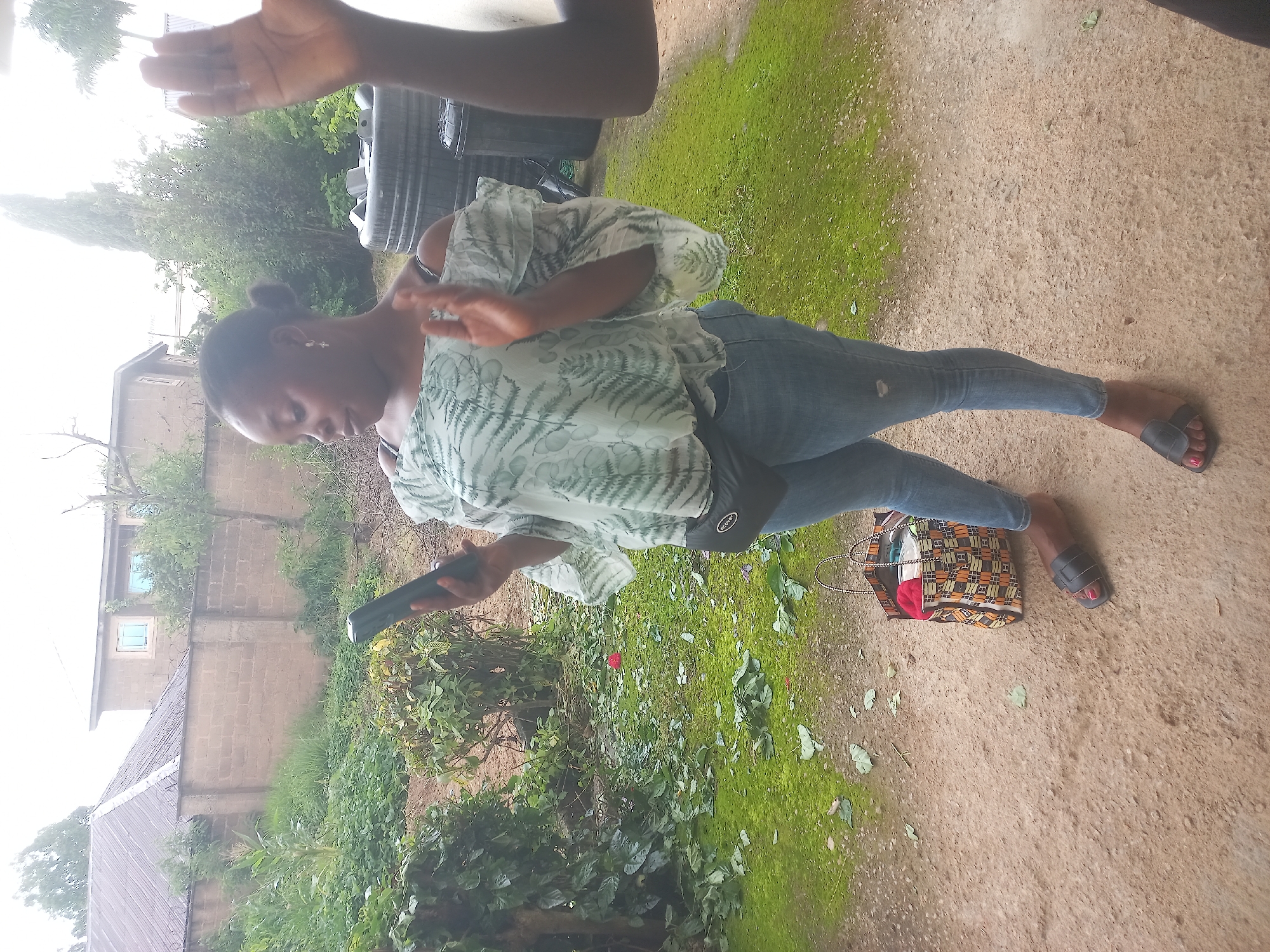 A full shot captures a Black woman standing outdoors and looking at her phone. She is wearing a light-colored, off-the-shoulder blouse with a fern-like pattern and light-wash jeans. A black fanny pack is around her waist. Her feet are adorned with black slide sandals. To her left, a colorful tote bag rests on the dirt ground. The background shows a fence, buildings, trees, and grass.