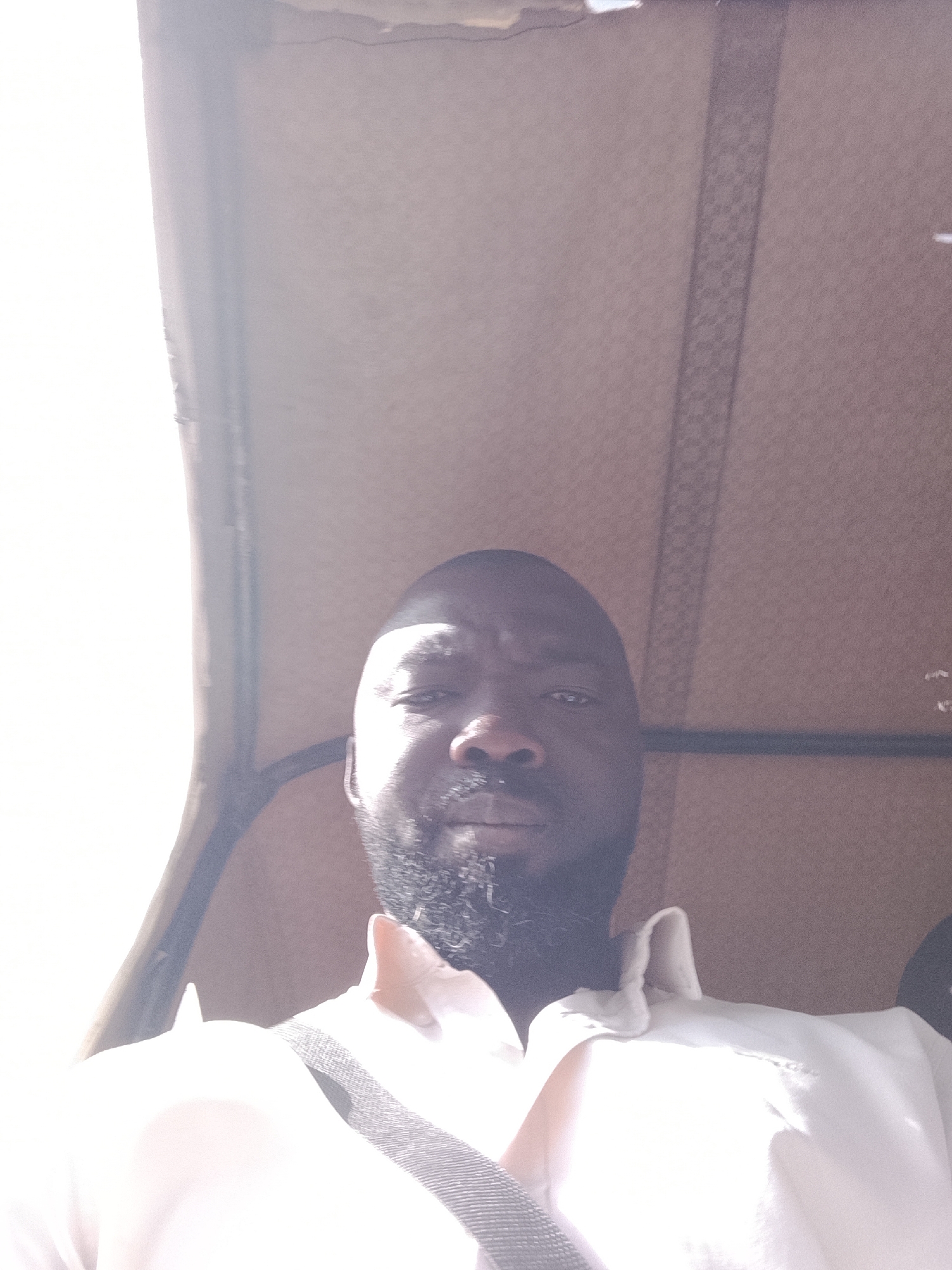 A dark-skinned man with a beard is taking a selfie inside a vehicle, possibly an auto-rickshaw, with a light-colored, patterned roof. He is wearing a white shirt and a strap across his chest. The location is assumed to be Jos, Nigeria.