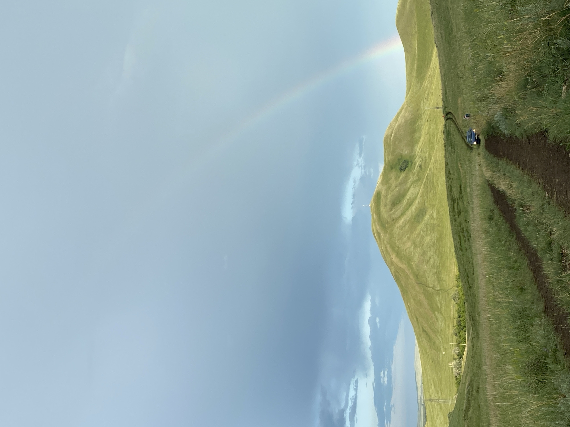 The image depicts a vast, rolling green landscape under a partly cloudy sky with a faint double rainbow visible. A dirt track winds its way through the tall grass, leading towards a prominent, rounded green hill in the middle ground. Atop this hill, a solitary white tower stands. Further along the track, a dark blue SUV is visible, its headlights on, with two people standing near it. The environment suggests a rural or natural setting, possibly on the outskirts of Верхнеозёрное, Russia, given the presence of the tower, which could be a telecommunications or observation tower. The overall mood is serene and tranquil, with the natural beauty of the landscape dominating the scene.