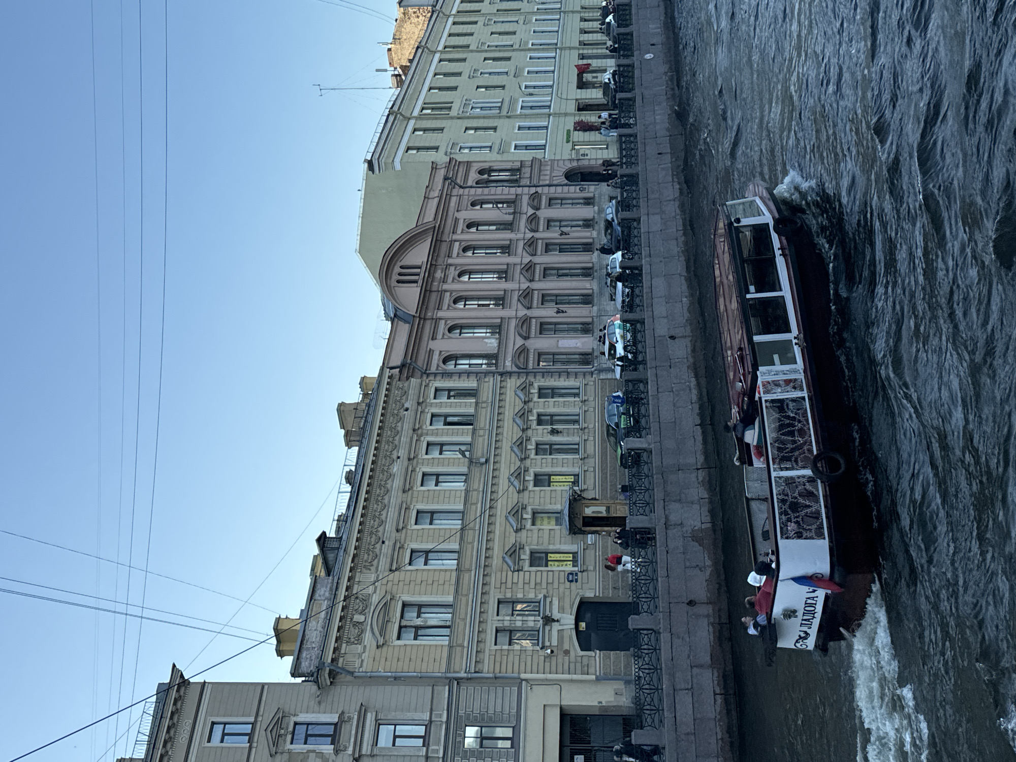 This is a photograph of a boat navigating a canal in Saint Petersburg, Russia. The boat, which appears to be a tour boat, is moving from left to right across the frame, leaving a wake in the dark, choppy water. The boat has "ЛАДОГА" (Ladoga) written on its stern, along with a Russian flag. Several people are visible on the boat, some seated and others standing.
Along the canal's embankment, there is a stone wall with a decorative metal railing. Behind the railing, a row of historic buildings lines the street. These buildings exhibit varied architectural styles and colors, including beige, pink, and pale green. They are multiple stories high and feature numerous windows. Some of the buildings have ornate architectural details.
On the sidewalk in front of the buildings, several people are walking or standing. Cars are parked along the street. Overhead, a clear blue sky is visible, with several power lines or telecommunication cables crisscrossing the upper portion of the image. The scene suggests a daytime setting, likely during fair weather.