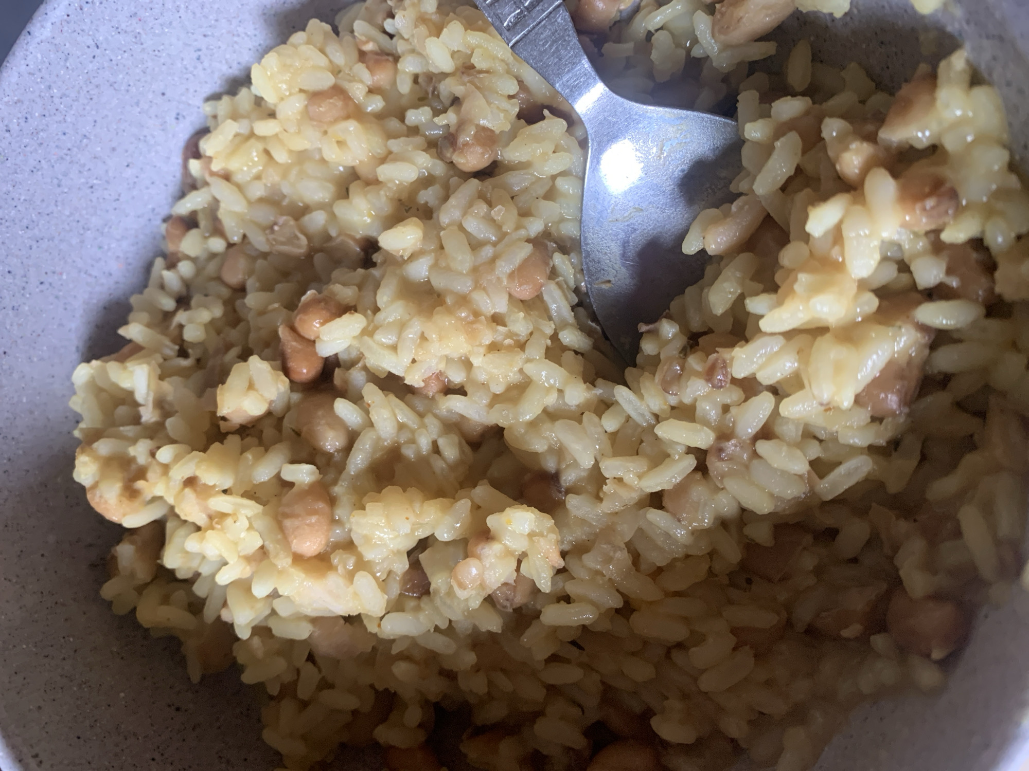 A close-up, eye-level shot shows a bowl of rice and beans. A silver spoon rests partially submerged in the mixture. The bowl is light gray with a speckled texture. The rice is a light yellow color, and the beans appear to be a light brown or pinkish hue. The dish is viewed from a top-down perspective in an indoor setting, with no discernible background or city information.