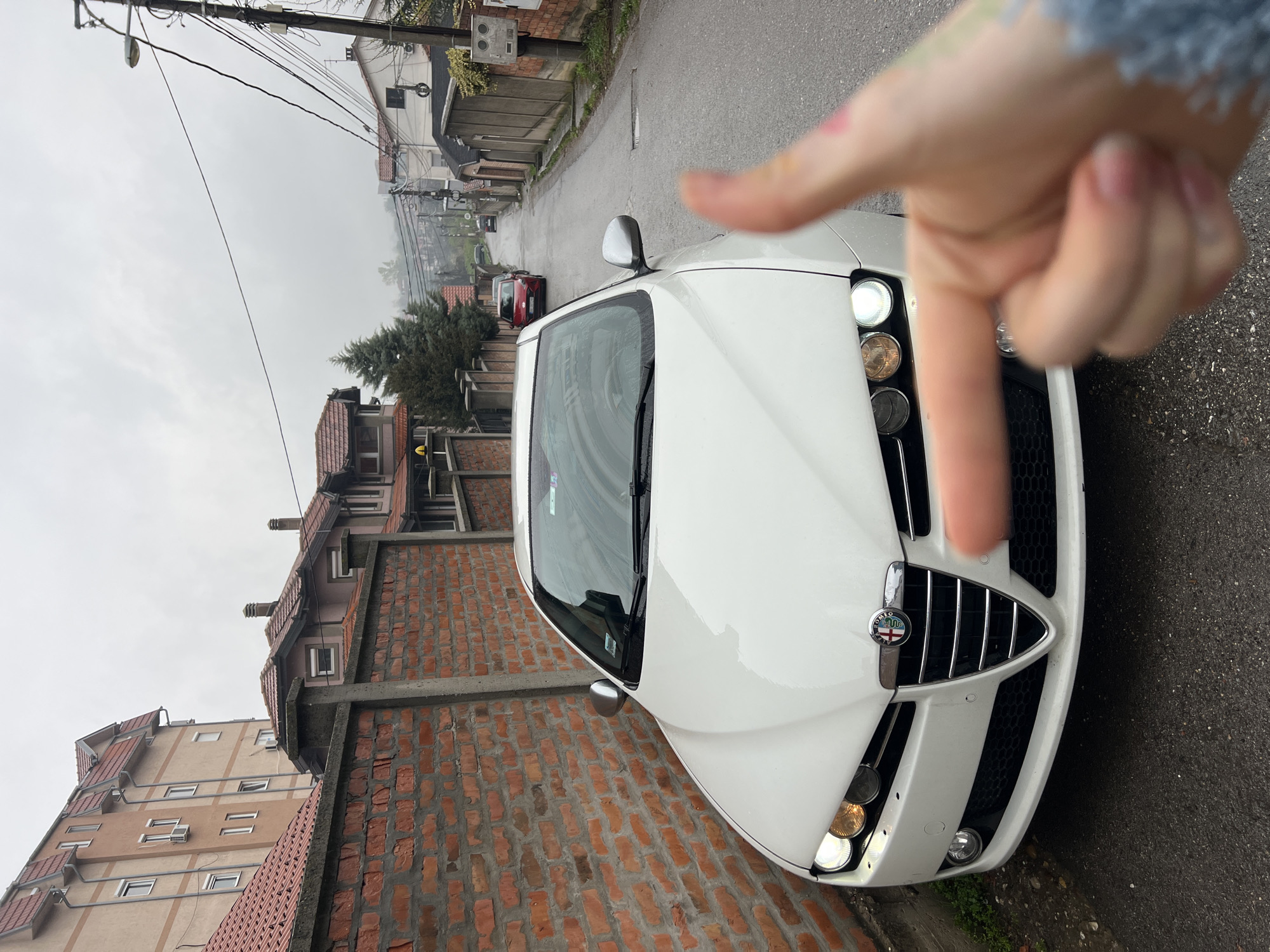 A white Alfa Romeo Brera is parked on a narrow street in Palilula Urban Municipality, Serbia. The car faces up the street. A person's hand and finger point towards the vehicle. The street is lined with brick buildings and houses. Overhead power lines are visible. The weather appears overcast and possibly drizzly.