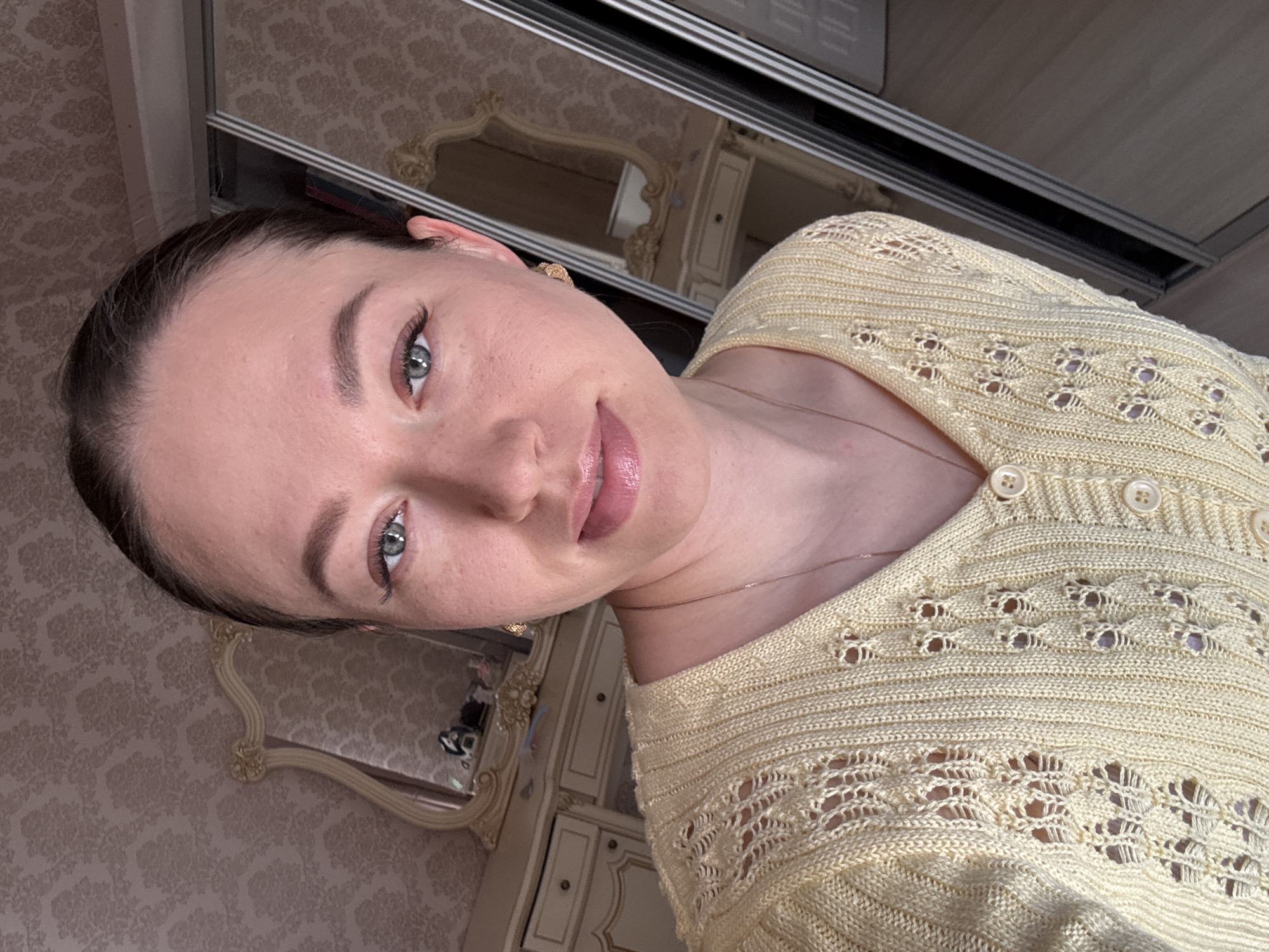 A woman is centered in the image, her face tilted towards the camera. She has fair skin, dark hair pulled back, and light-colored eyes. She is wearing a light yellow knit cardigan with decorative openwork. A thin gold necklace is visible around her neck.
Behind her, a mirror reflects a portion of a room, including what appears to be decorative wallpaper and furniture with ornate details. Above her is a sliding door track, possibly of a closet.
Given the prompt's mention of Ферма, Russia, the location is likely a private residence in that city.