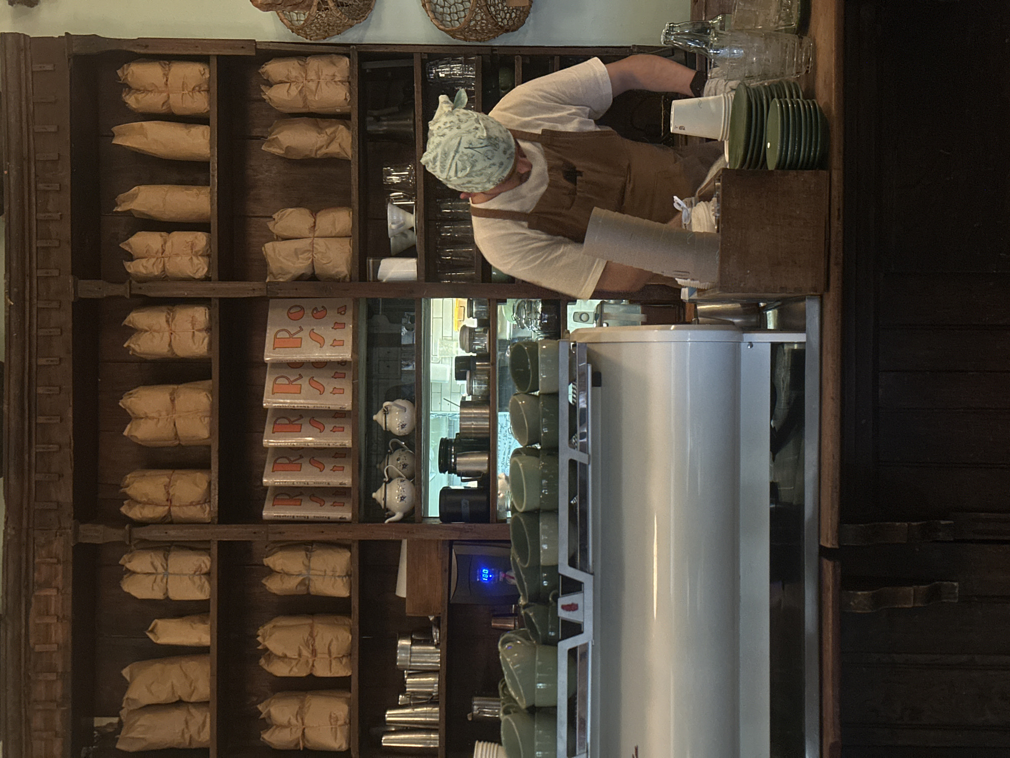 The image depicts the interior of a shop, likely a coffee shop, in Mexico City, Mexico. A worker wearing a head covering and an apron is positioned behind a counter. The background features a wooden shelf filled with paper-wrapped bundles, books, teapots, and metal containers. A large coffee machine is visible in the foreground. There are also stacks of green plates and clear glassware on the counter.