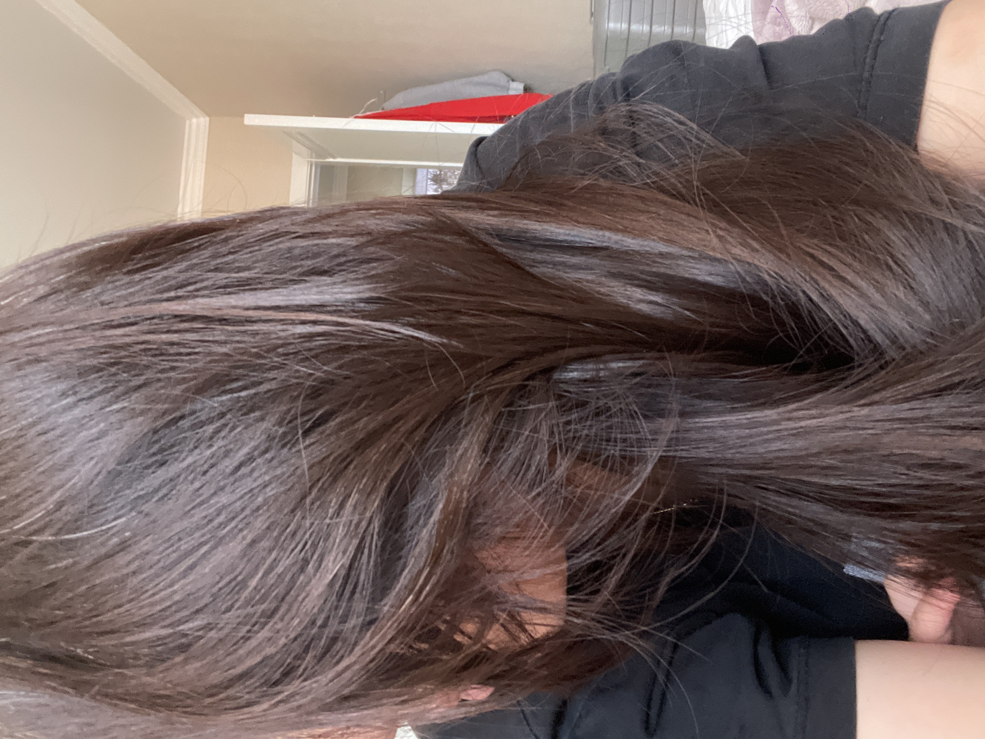 The image shows a close-up of a person with long, dark brown hair lying on a black shirt. The background is out of focus and shows a room with a beige wall and a glimpse of a white shelf with some items on it. The person's face is partially visible and obscured by their hair. The location is not identifiable as Kokshetau, Kazakhstan based on the image alone. There are no discernible actions or interactions taking place in the image.