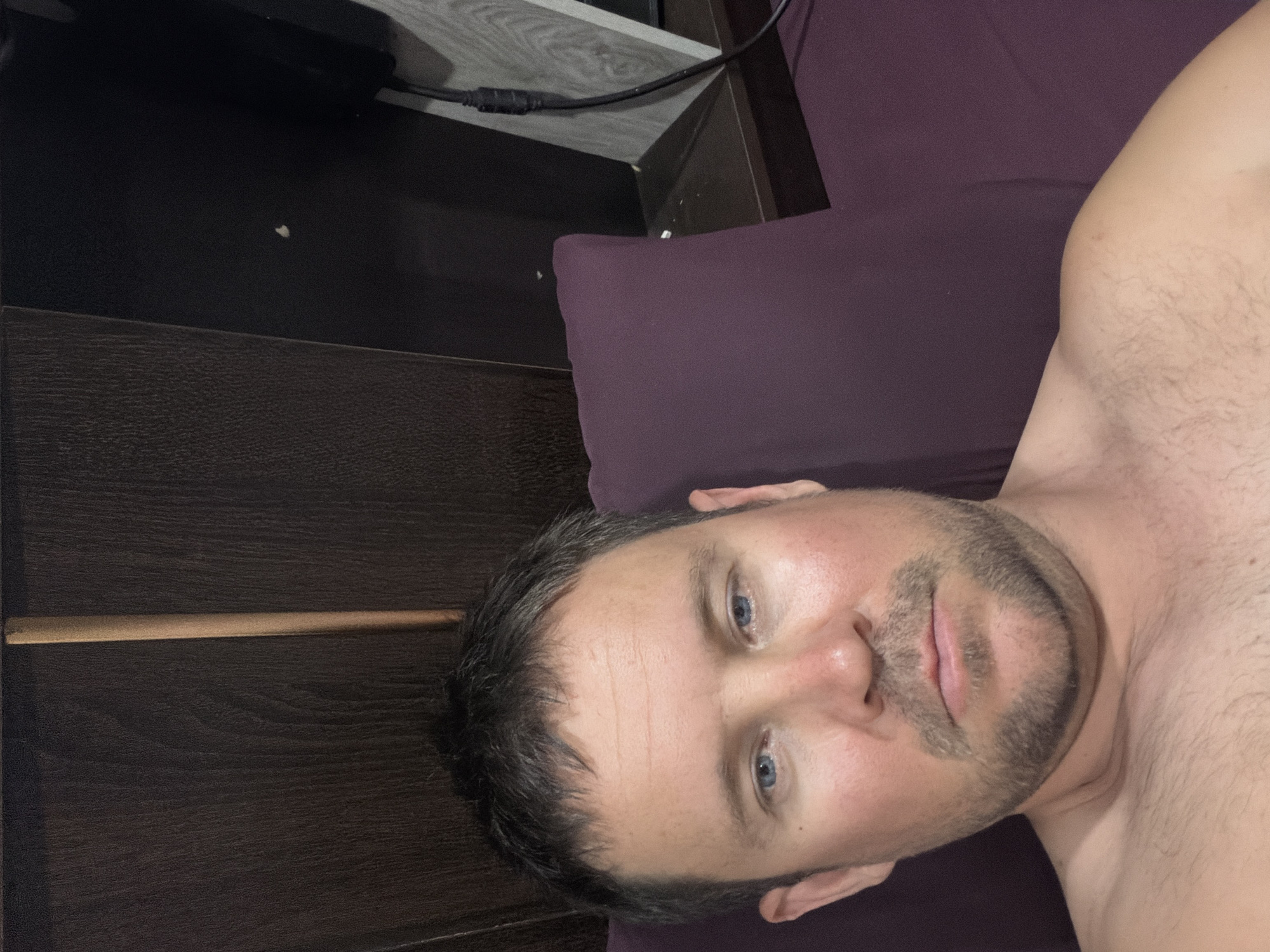 A man is lying down on a purple pillow in what appears to be a bedroom. The man has light skin, short brown hair, and blue eyes. He is wearing a dark t-shirt and has stubble on his face. The room has dark wood paneling on the wall behind him, and a shelf with some electronic equipment. The man is looking at the camera. The image is a close-up of the man's face and upper body. The lighting is dim.