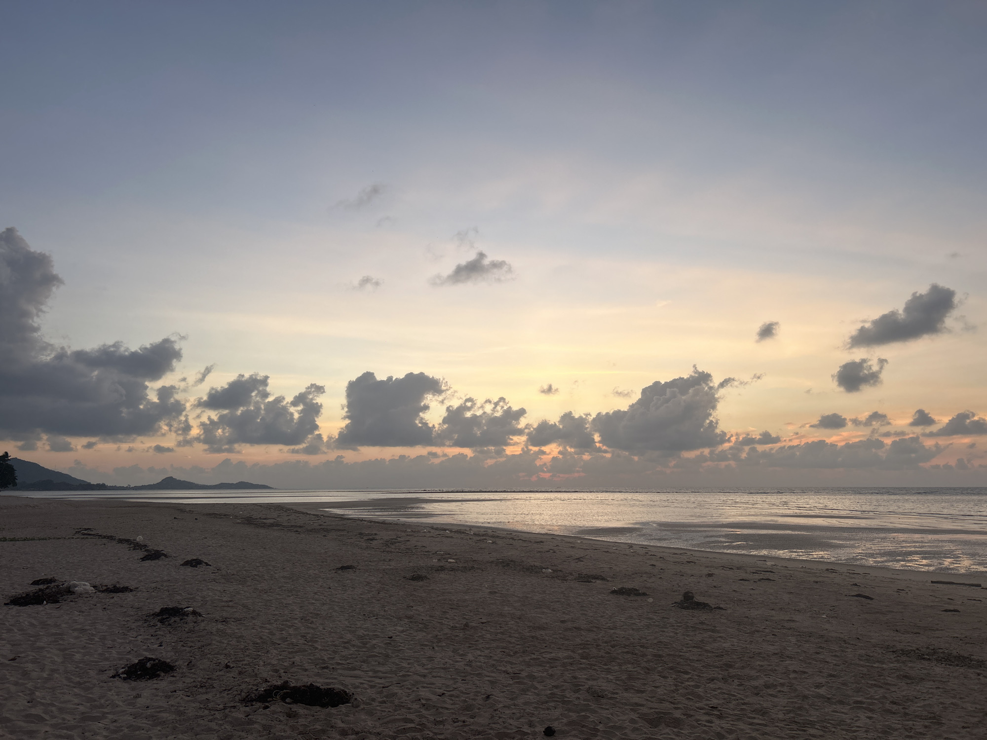 This media file captures a serene coastal landscape at either sunrise or sunset, presenting a wide, tranquil beach scene. The setting is Baan Harn, Thailand.
The sky exhibits a gentle gradient of colors, transitioning from soft pale yellow and orange hues near the horizon to a lighter blue and muted grey overhead. Scattered across the sky are numerous clouds, appearing dark grey in silhouette against the soft light, with some larger, more defined cumulus formations, particularly on the left side and along the horizon.
The foreground is dominated by a wide expanse of dark, damp sand, stretching from the bottom of the frame towards the water. The sand appears undisturbed except for several small, dark piles of what resembles seaweed or natural debris, loosely scattered across the beach. No people or human structures are visible on the beach itself.
Further back, the calm waters of the sea or a large gulf gently meet the shore. The water surface is smooth, reflecting the soft, warm light of the sky, creating shimmering patterns. The low tide reveals vast areas of wet sand and shallow water along the shoreline. In the distance, a subtle horizon line separates the water from low-lying hills or mountains, visible on the left side of the frame, adding depth to the scene.
The overall impression is one of peaceful solitude and natural beauty, characteristic of a quiet coastal morning or evening. There is no visible text in the image.