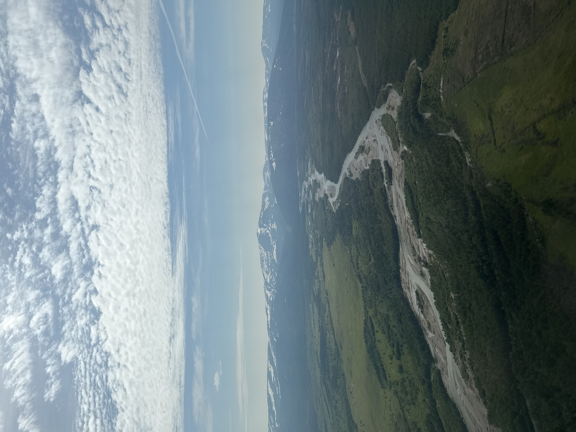 An aerial view of a vast, sparsely populated landscape with a wide, meandering river flowing through it. The riverbed appears to be sandy or gravelly, with multiple braided channels. Lush green forests and open fields surround the river, transitioning into a mountainous region in the distance, capped with snow. The sky is partly cloudy with scattered white clouds and a clear blue sky. The scene depicts a remote natural environment, likely in the wilderness of Stekolny, Russia, with no signs of human activity other than a faint contrail in the sky.