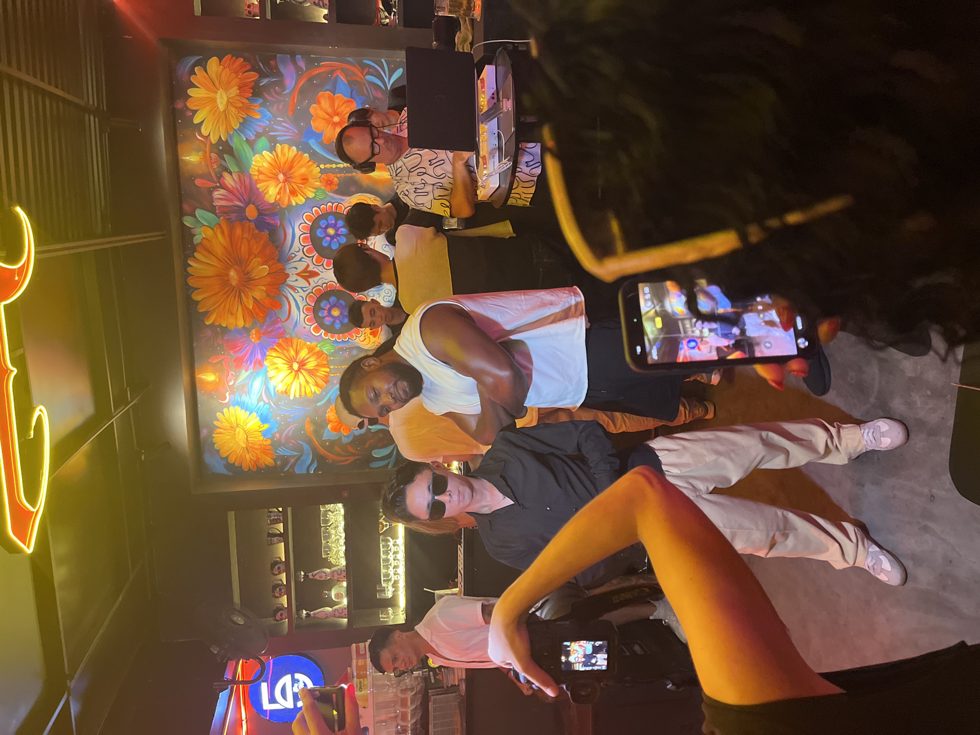 The image captures a group of individuals posing for pictures in what appears to be a bar or entertainment venue in Đà Nẵng, Vietnam. The location is identifiable by the neon sign with Vietnamese characters on the left side of the image. The background features colorful artwork, including floral designs.
Several people are visible, some posing directly for the camera, while others are taking pictures with their phones. A DJ with headphones is seen near a laptop and sound equipment, suggesting the venue may feature live music or DJ sets.
The focus is on the group of people posing for pictures, with individuals wearing various styles of clothing, including casual shirts, pants, and sunglasses. The environment is dimly lit, creating an atmosphere suitable for a bar or nightclub setting.