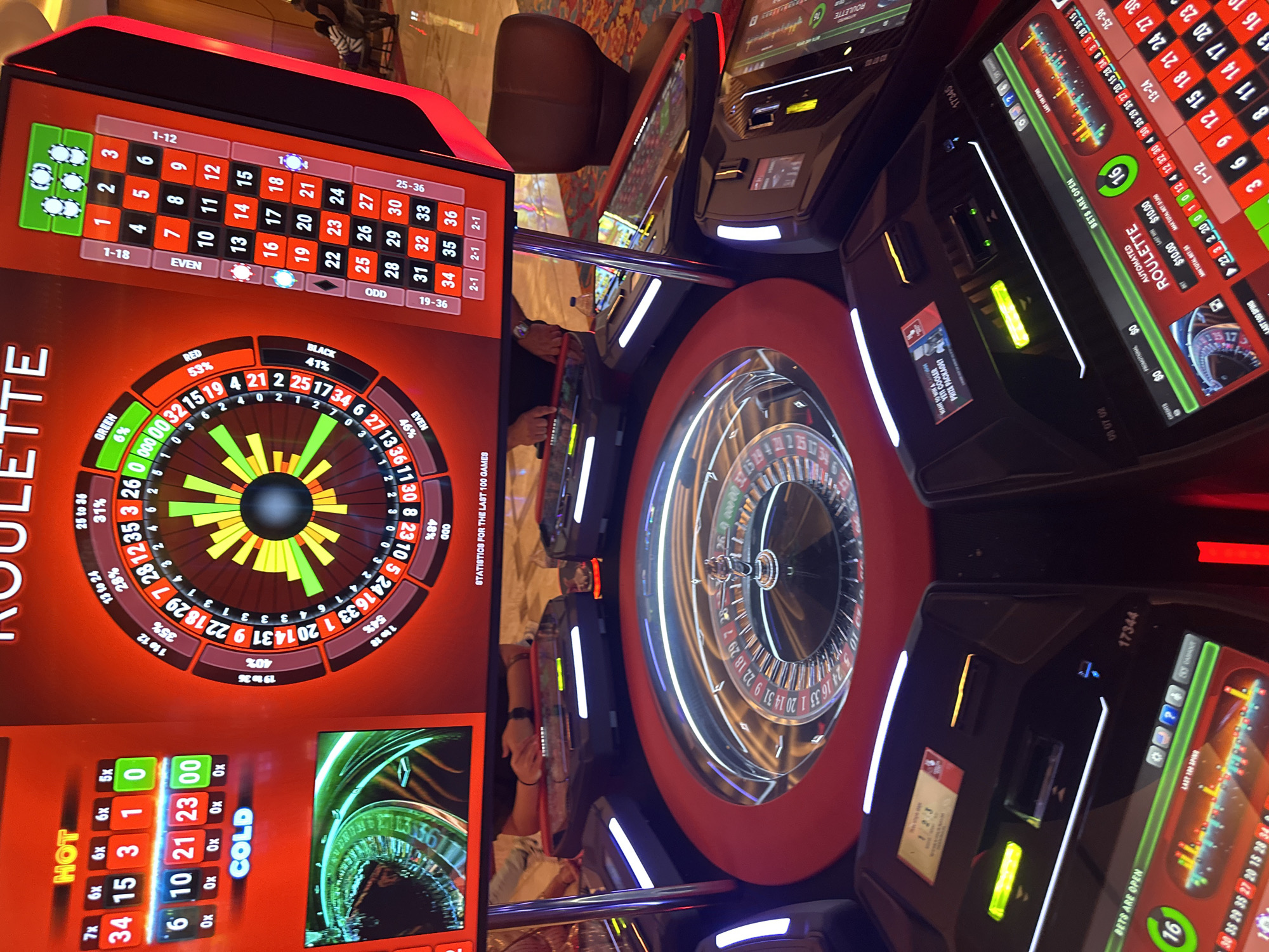 Here is a description of the image:
**Location:** The image appears to be taken in a casino, likely at the Seminole Hard Rock Hotel & Casino on the Hollywood Seminole Reservation, Florida, USA.
**Subjects and Objects:** The primary focus is on several automated roulette machines. These machines feature digital displays showing the roulette wheel, betting options, game statistics, and "hot" and "cold" numbers. The physical roulette wheels themselves are visible within the circular machines. A chair is partially visible in the background. The reflections in the glass surfaces of the machines suggest a brightly lit environment and possibly other casino patrons nearby.
**Details:**
* The digital displays of the roulette machines show various betting options and odds.
* The "hot" and "cold" number indicators display which numbers have recently appeared more or less frequently.
* The roulette wheels are in motion, with the ball visible in some of the wheels.
**Environment:** The casino environment is evident from the design of the machines, the lighting, and the overall ambiance suggested by the reflective surfaces. The details suggest a high-end casino setting.