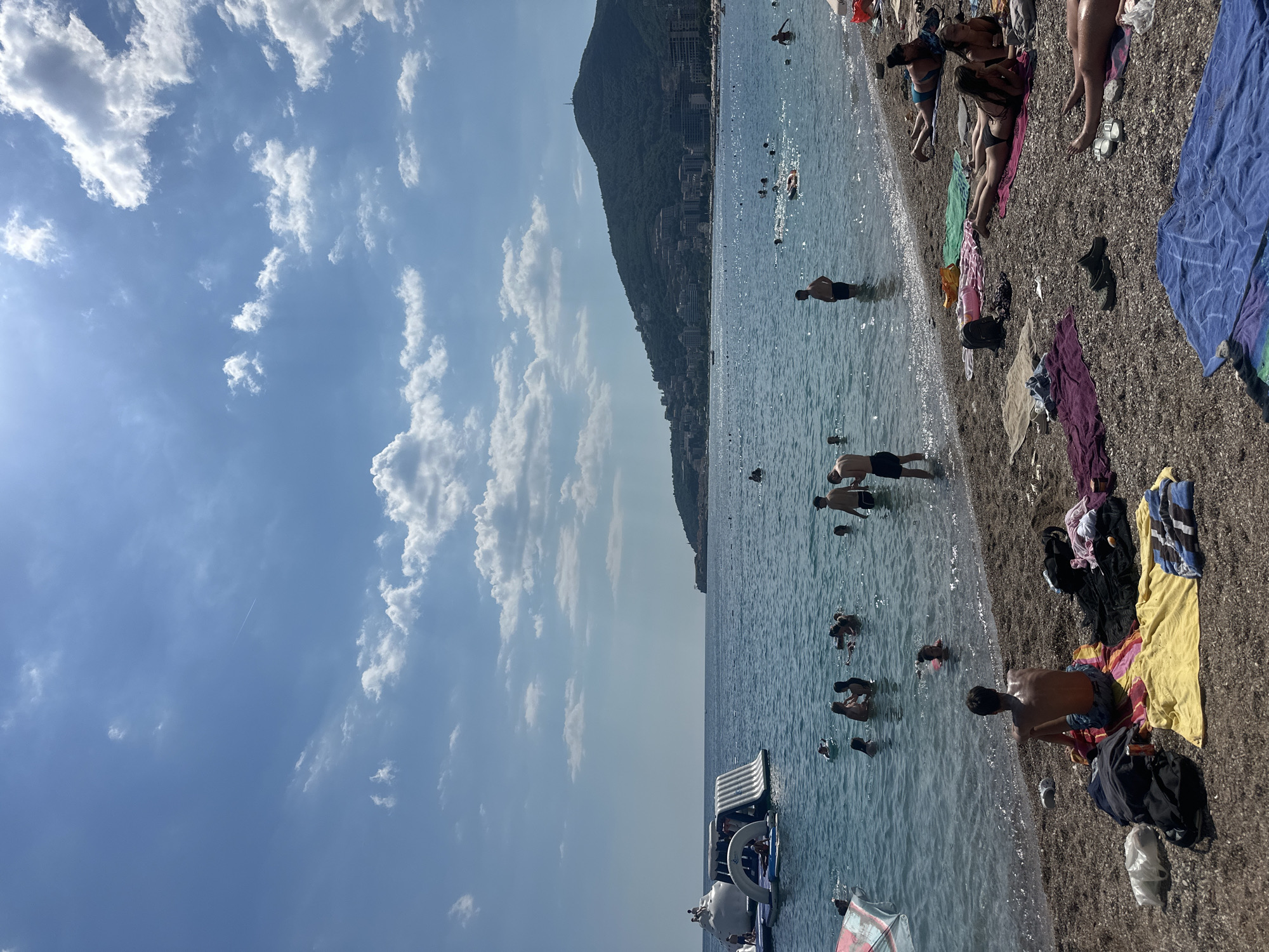 The image depicts a lively scene at a pebble beach in Budva, Montenegro, under a clear blue sky dotted with fluffy white clouds. The turquoise water of the Adriatic Sea is populated with numerous people swimming and enjoying water activities. In the distance, a large inflatable water park structure is visible on the sea. The shoreline is lined with people relaxing on towels, sunbathing, and wading in the shallow water. Some individuals are sitting on the beach, while others are lying down, all engaged in typical beach leisure. The backdrop features a verdant, hilly coastline with buildings cascading down its slopes, characteristic of Budva's urban landscape. The overall atmosphere is one of a vibrant summer day at a popular coastal destination.
