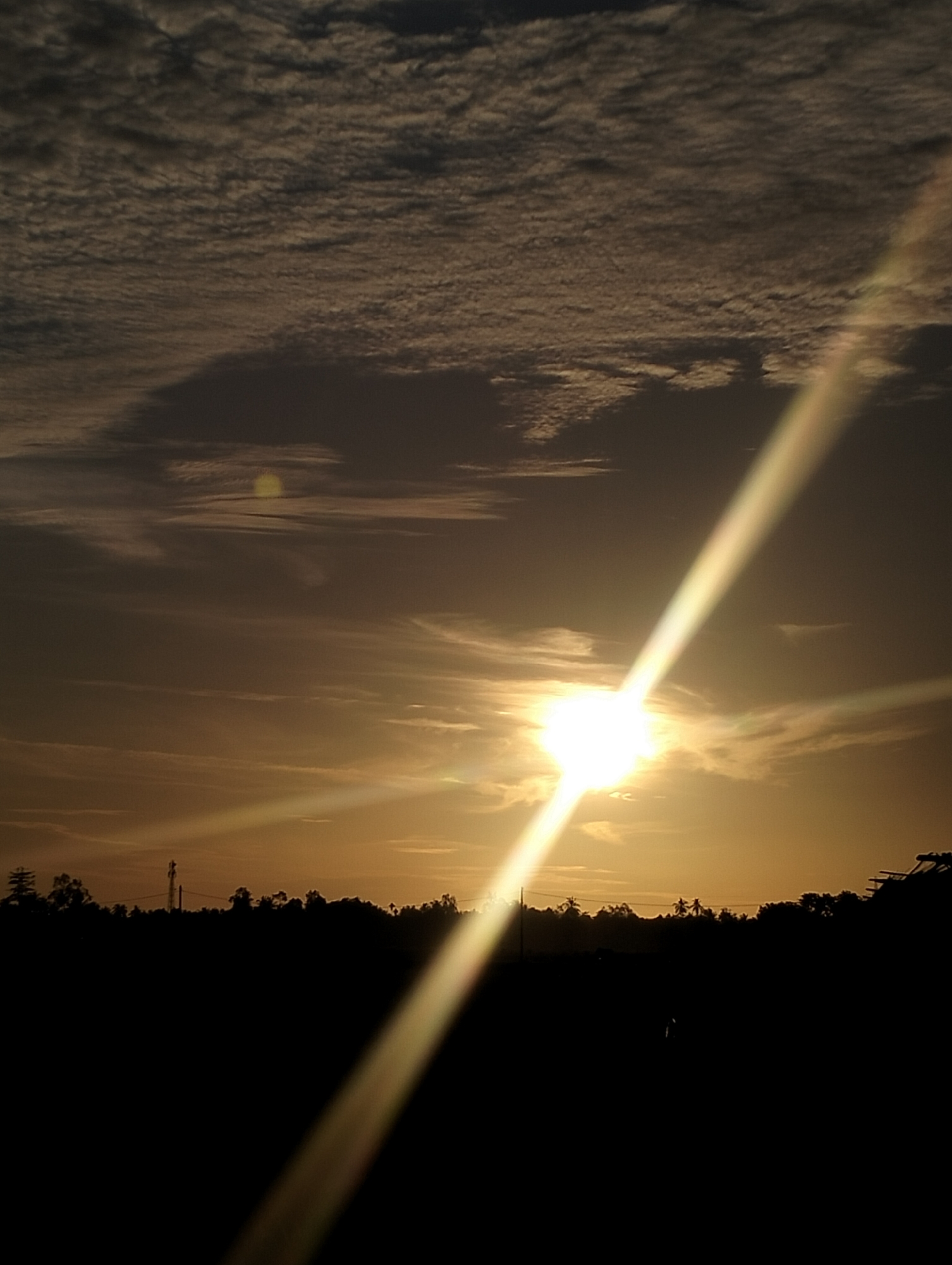 The image depicts a sunset in Kuta Pungwa, Indonesia. The sun is low on the horizon, obscured by thin clouds and casting a bright glare with defined light rays. Silhouetted trees and foliage form a dark, uneven foreground. Overhead, the sky shows a mix of clouds with varying density and texture. A telecommunications tower is visible in the tree line.