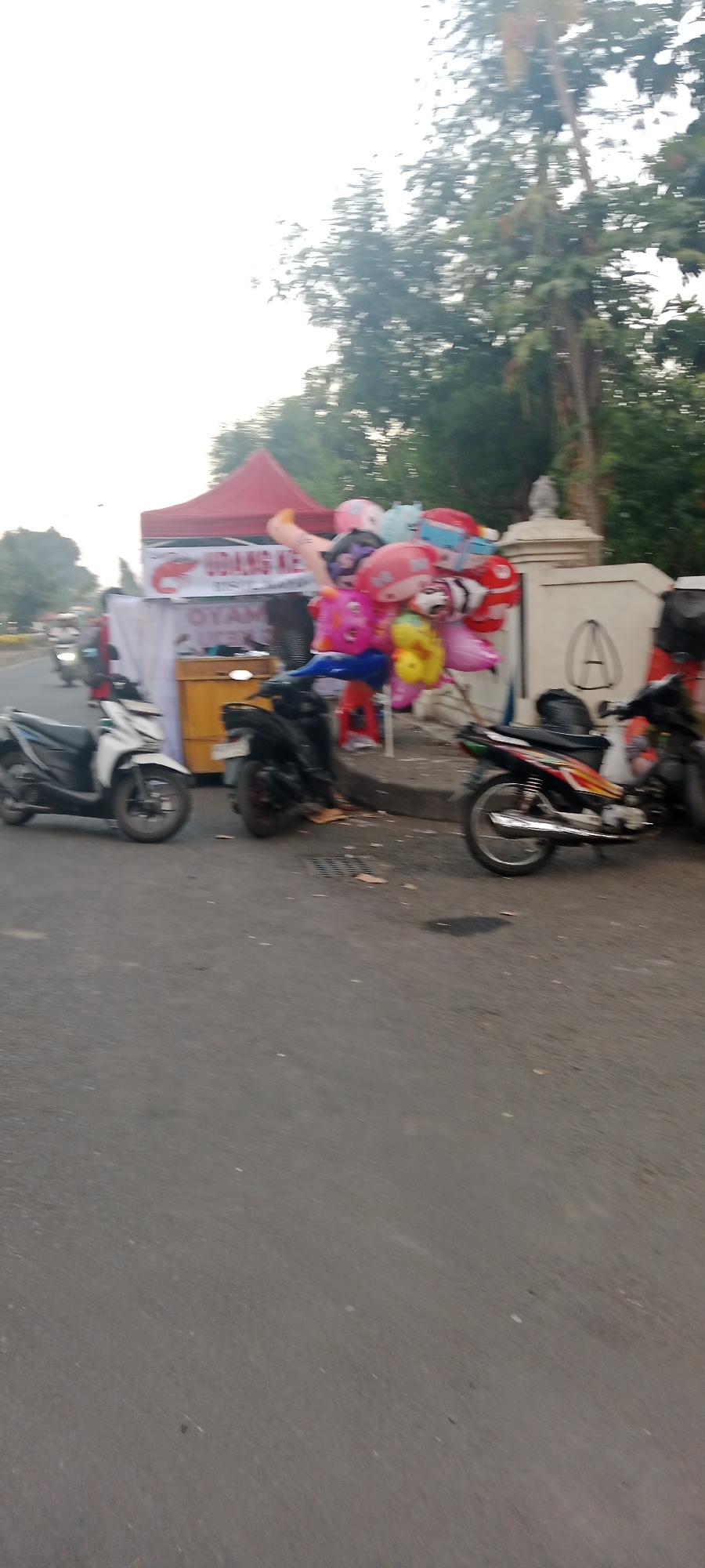 In Ngemplak, Indonesia, this image captures a street scene with a red tent set up on the side of the road. A cart is visible under the tent, and a variety of colorful balloons are displayed. Two motorcycles are parked near the tent, and the road surface is asphalt. Trees line the background, and the sky is overcast. There are no people clearly visible in the image, but it appears to be a commercial area.