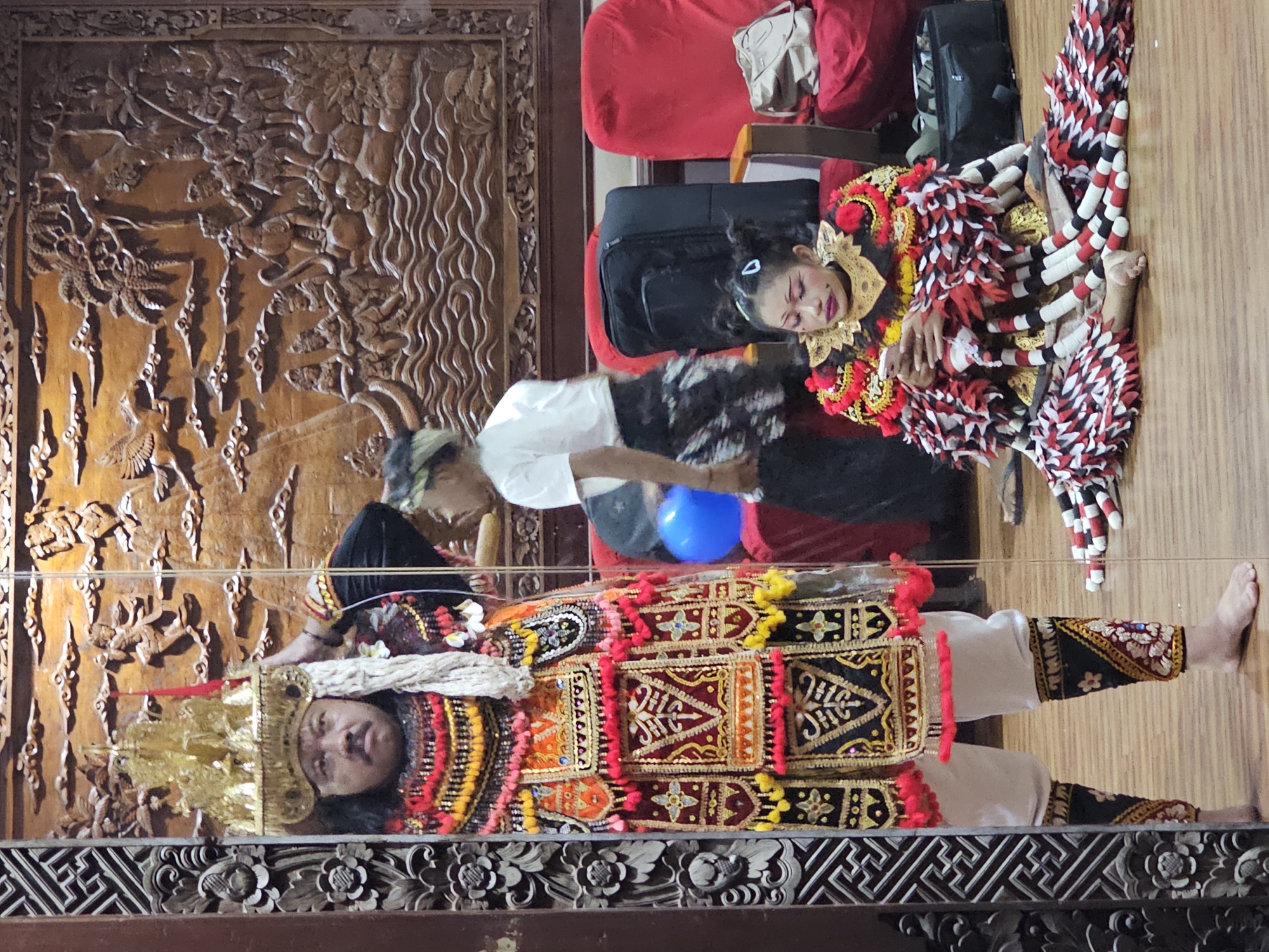 In Denpasar, Indonesia, a cultural performance is underway, reflected in a large mirror that dominates the foreground. A man, adorned in an elaborate golden headdress and a vibrantly colored, tasselled costume featuring intricate embroidery, stands reflected in the mirror, his hand touching his headdress. Beside him, a young girl, also in traditional attire with a distinctive red and white feathered skirt and striking makeup, sits on the floor, looking at her phone. In the background, a person in a white shirt and dark skirt stands near a blue balloon and a large black suitcase, with red chairs visible. The scene is set against a backdrop of a large, ornate wooden carving depicting a mythological scene with figures and stylized clouds. The flooring is polished wood.