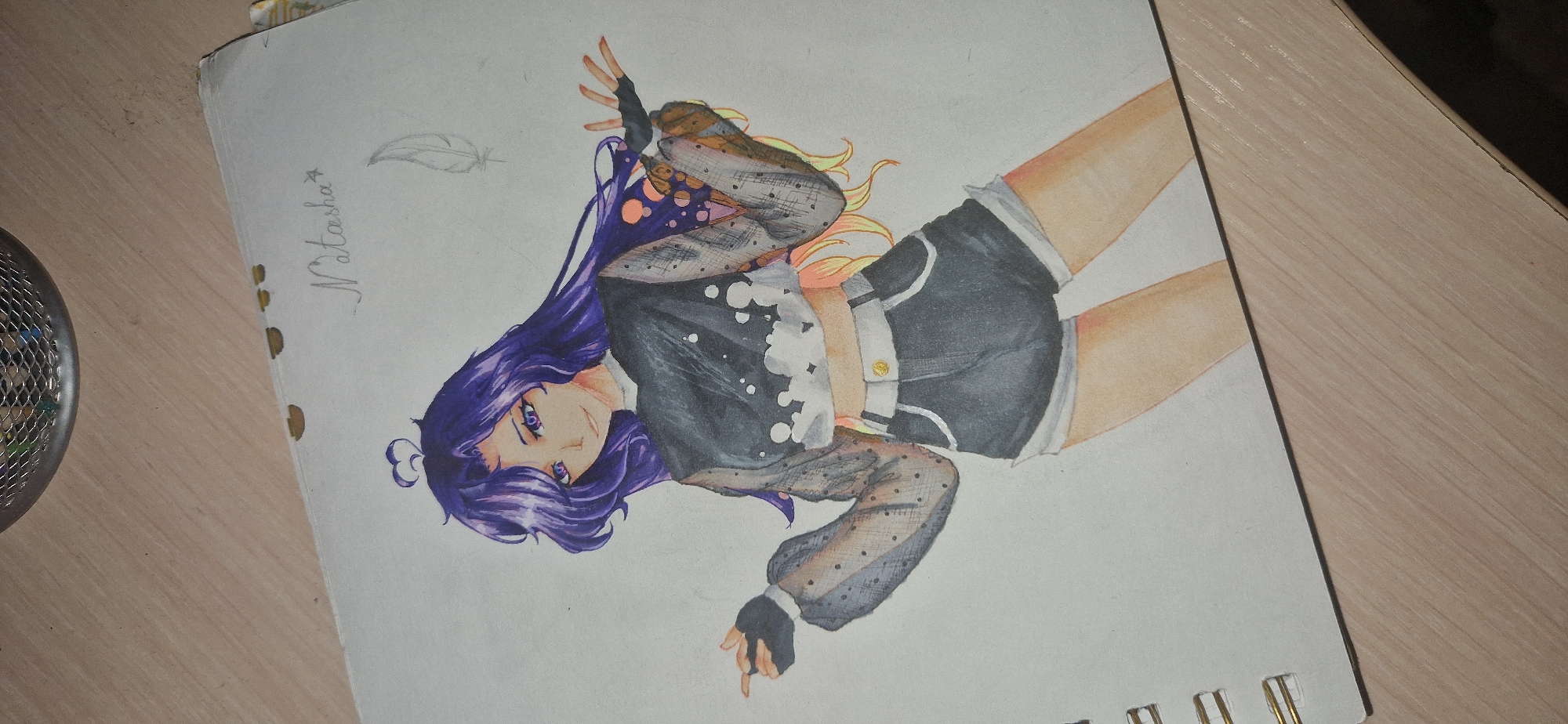 A drawing on paper depicts an anime-style female character with long, wavy purple hair and purple eyes, wearing a black mesh crop top with white splatter-like accents and black denim shorts. The character is striking a dynamic pose with one hand raised. The drawing is signed "Natasha" in the upper right corner, accompanied by a small feather sketch. The paper is part of a spiral-bound notebook and is placed on a light-colored wooden surface. There is no indication of the city of Krasnoobsk, Russia, in this image.