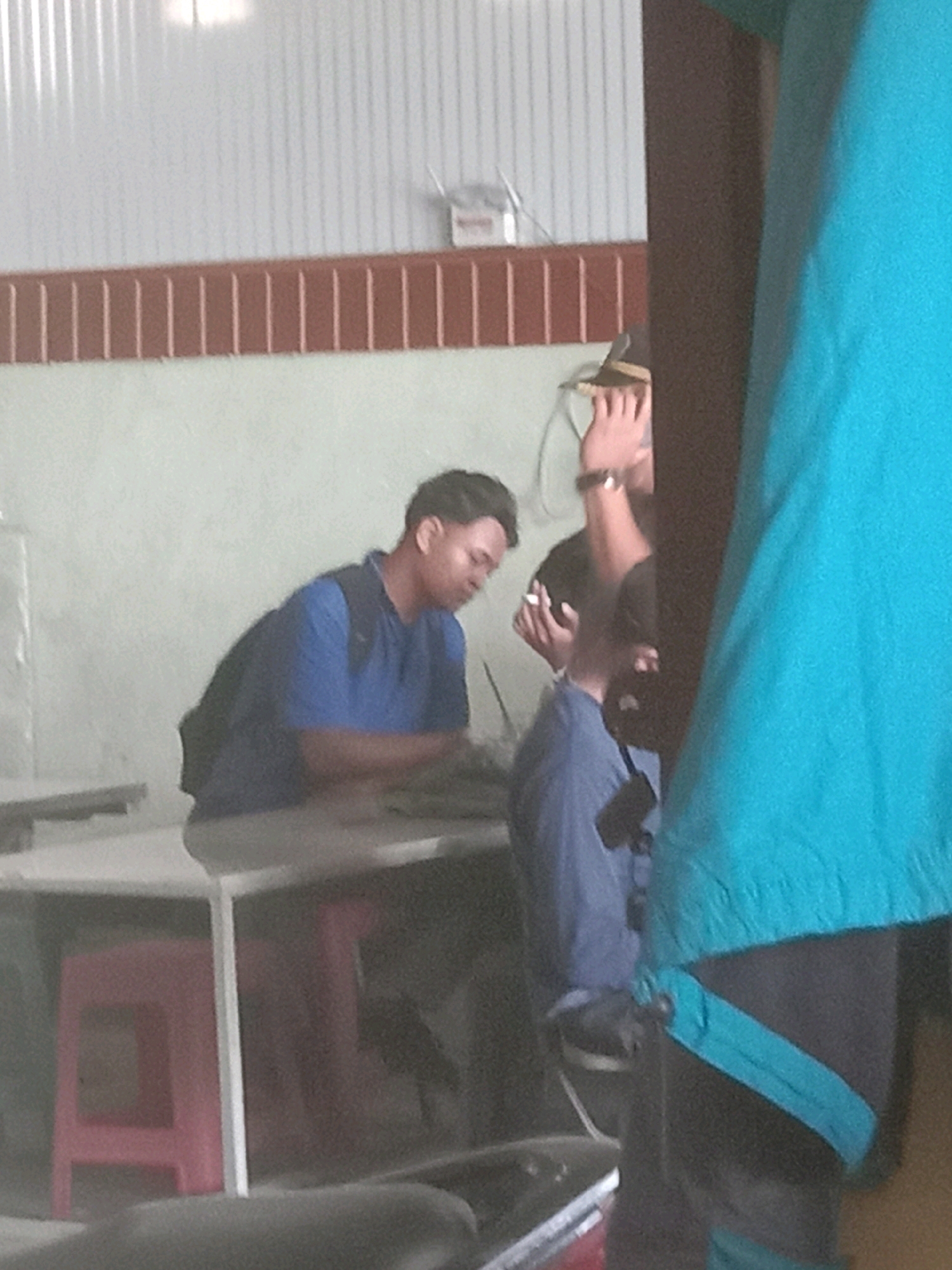 A medium shot captures three individuals in an indoor setting, possibly a restaurant or communal area, with corrugated metal walls and a light-colored wall in the background. The lighting is ambient. On the left, a person wearing a blue polo shirt and a backpack is seated at a table, looking downwards. In the center, another person, partly obscured by the person on the right, is holding a cigarette to their mouth and looking to the left. The person on the right is mostly out of frame, wearing a bright teal garment that drapes over their shoulder and torso. Red stools are visible beneath a table to the left. The scene suggests a casual gathering or public space. The location is identified as Plosowahyu, Indonesia.