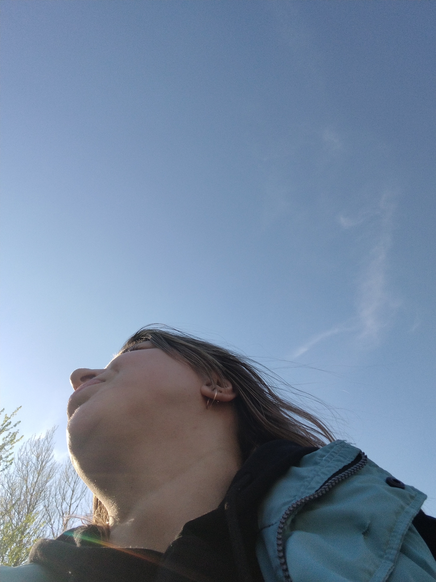 The image is a low-angle shot of a person looking up at a clear blue sky with a wispy cloud formation in the upper right. The person has light skin and wears a teal jacket over a dark-colored shirt. The background includes bare trees. The location is unknown and no information can determine that it is located in Шебунино, Russia.