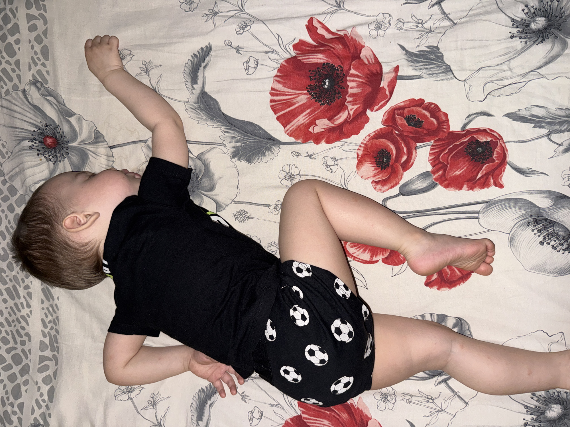 A child lies on a floral-patterned bedspread. The child is wearing a black t-shirt and black shorts with white soccer balls. The child's left arm is raised above their head. The child's face is turned away from the camera. The location is unknown.