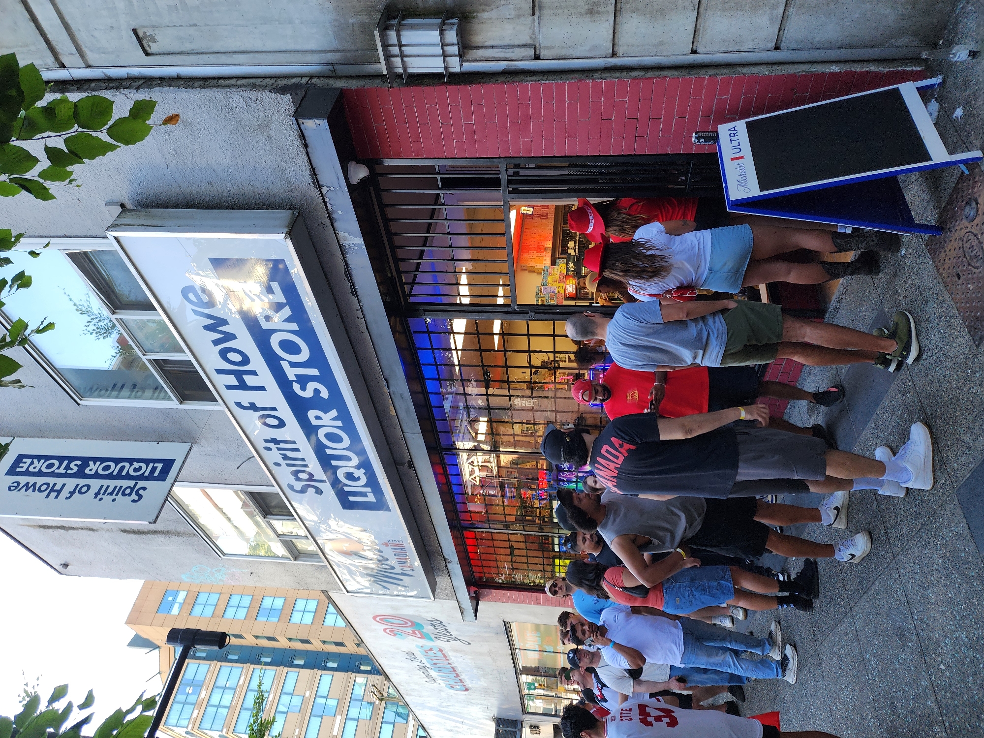 A group of people are standing in line outside a liquor store called "Spirit of Howe Liquor Store." The building is made of grey concrete with red brick accents. There are large windows at the front of the store, which are barred from the outside. Inside the store, shelves are stocked with bottles. The street is paved with grey asphalt, and there are some leafy trees to the right of the building. One person in line is wearing a t-shirt that says "CANADA." A blue and white A-frame sign for Michelob Ultra is standing on the sidewalk. The sky is a bright, overcast grey.