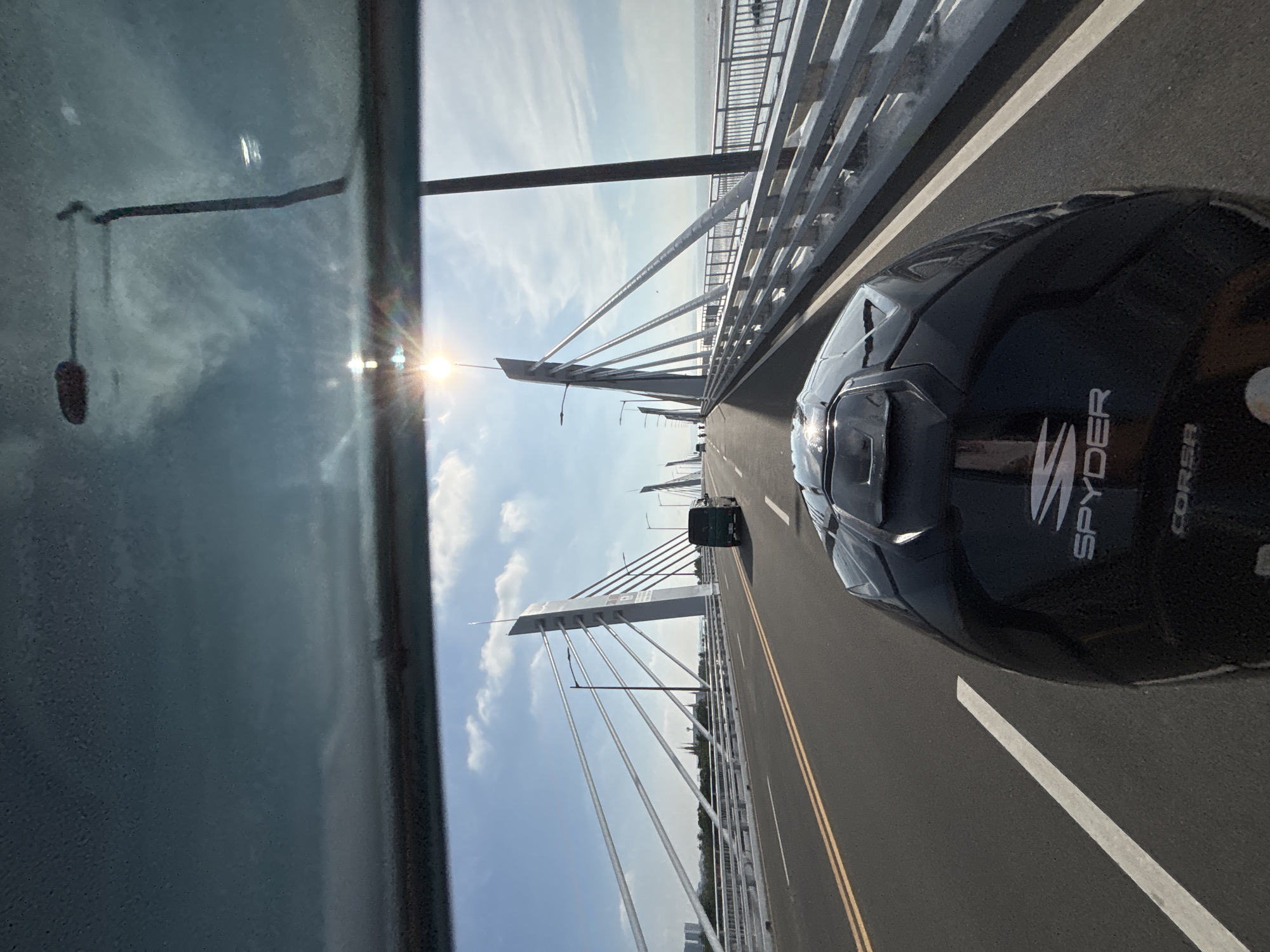 The image shows a first-person view of someone riding a motorcycle on a bridge. The motorcycle is black with "SPYDER" and "CORSA" logos visible on the helmet. The bridge has multiple lanes and is characterized by tall, modern pylons with cables supporting the deck. The sun is shining brightly, casting lens flare effects on the windshield. In the background, there are other vehicles, including a dark green van. The bridge extends over a body of water, with a railing visible along the side. Distant buildings and a hazy skyline are visible on the left. This scene is consistent with the iconic Francisco Bangoy International Airport bridge in Davao City, Philippines, which connects the mainland to the airport island and is known for its distinctive architecture and waterside location.