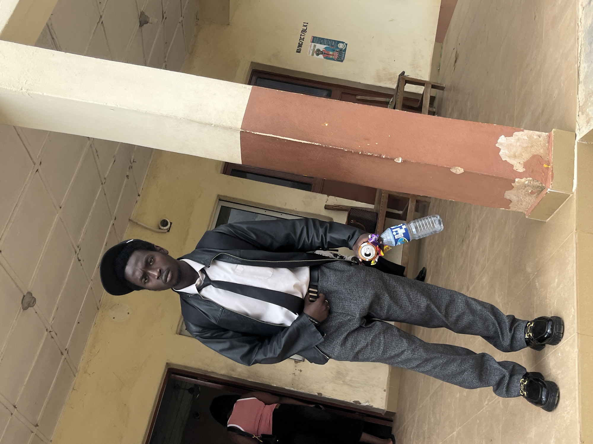 A young Black male stands on a tiled patio in front of a building with a light yellow facade and a large concrete pillar painted brown and white. He is wearing a black baseball cap, a black leather jacket, a white dress shirt, a black tie, grey patterned trousers, and black patent leather loafers with gold accents. He is holding a can and a plastic bottle of water in his right hand. Behind him, a woman in a pink top and black skirt is seen exiting a doorway. The building appears to be a school or an office, indicated by a poster on the wall bearing text that includes "UI/NC/ICT/BLK1" and a portrait of a man in an orange vest. The overall environment suggests a warm climate. The image does not contain enough information to definitively place it in Jos, Nigeria.