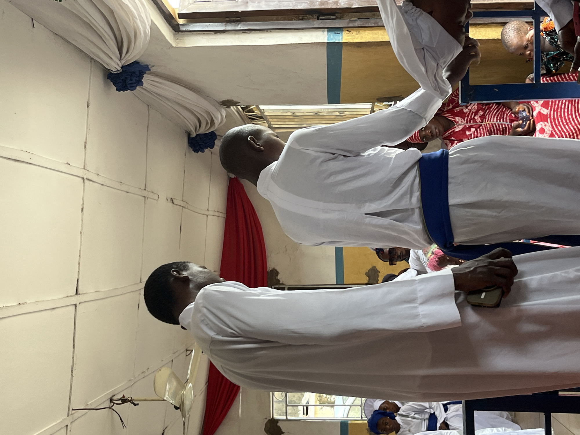 Two young men in white robes stand facing away from the camera in what appears to be an indoor gathering. The man on the left wears a plain white robe and holds a small, light-colored object in his right hand, partially obscured by his robe. The man on the right wears a white robe with a blue sash around his waist and is leaning slightly forward, with his head bowed. Behind them, a group of people are seated, including children. One young girl in the background wears a blue headscarf and a white garment with a blue sash. Another person, likely a woman, is seated and wears a red and white patterned outfit. The environment looks like a community hall or place of worship, with a visible ceiling fan, draped fabric in red and white, and yellow and blue painted walls. There is a metal-barred window to the right, through which daylight enters. The overall atmosphere suggests a ceremony or formal gathering.