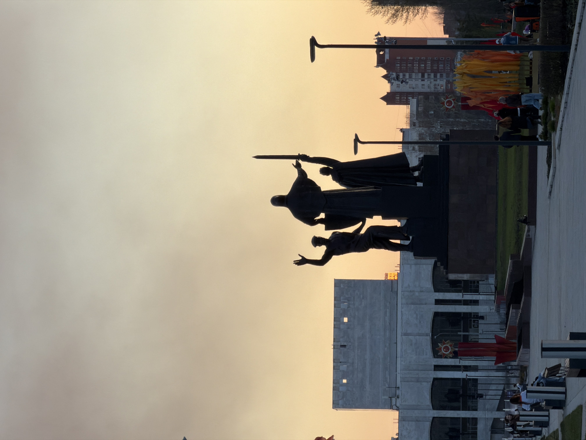 The image depicts a monument in Perm, Russia, silhouetted against a light-colored sky. The monument features three figures: one holding a sword aloft, another behind him, and a third figure reaching upward. Below the monument is a building with windows decorated with red stars and rocket-shaped objects. To the right of the building, there is a walkway with benches where people are sitting. Street lamps are visible above the monument, along with other buildings and colorful flags in the background. The scene appears to be taken at dusk or dawn.
