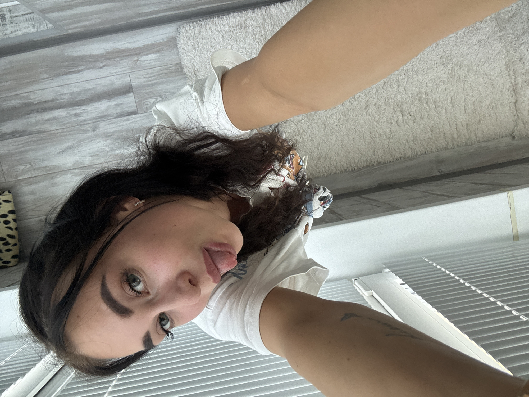 A young adult female is depicted in a selfie, taken from a slightly elevated angle. She has dark brown hair, prominent eyebrows, and light-colored eyes (appears hazel or light green). Her skin is fair, with visible freckles across her nose and cheeks. She is wearing a white t-shirt, part of which features a faded graphic design on her chest. A small, sparkling stud earring is visible in her left earlobe. A faint, indistinct tattoo is present on her left forearm. She is looking directly at the camera with a playful expression, extending her tongue outwards.
The setting is indoors, resembling a casual residential space. Bright, diffuse natural light illuminates the scene, likely originating from a window on the viewer's left side. Vertical blinds, partially open, are visible in the background on the left. The floor consists of light gray, wood-effect laminate or plank flooring, transitioning into a textured, light-colored (off-white/beige) shaggy rug in the lower right portion of the frame. A small segment of a patterned object, possibly a cushion or blanket with a dark animal print, is visible in the upper left corner of the background.
The image does not contain any visual identifiers or markers that establish a specific geographic location, including the city of Chaikovsky, Russia.