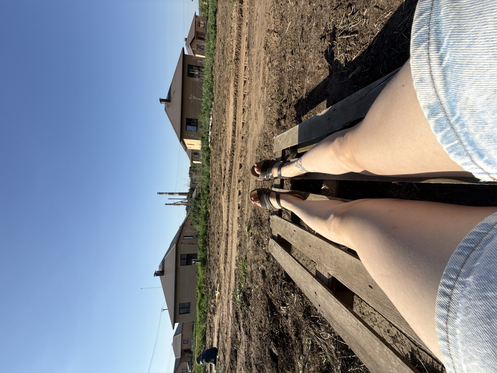 A horizontal shot features a low-angle perspective of a person sitting on a wooden pallet. The individual's legs, adorned with brown sandals, are visible from the knees down and are framed by the edge of light-wash denim shorts. The pallet is positioned on uneven, muddy ground interspersed with patches of grass and weeds.
In the background, brick houses with dark-colored roofs stand along a dirt road. In the distance, industrial smokestacks pierce a clear, blue sky.
The scene is in Chaikovsky, Russia.