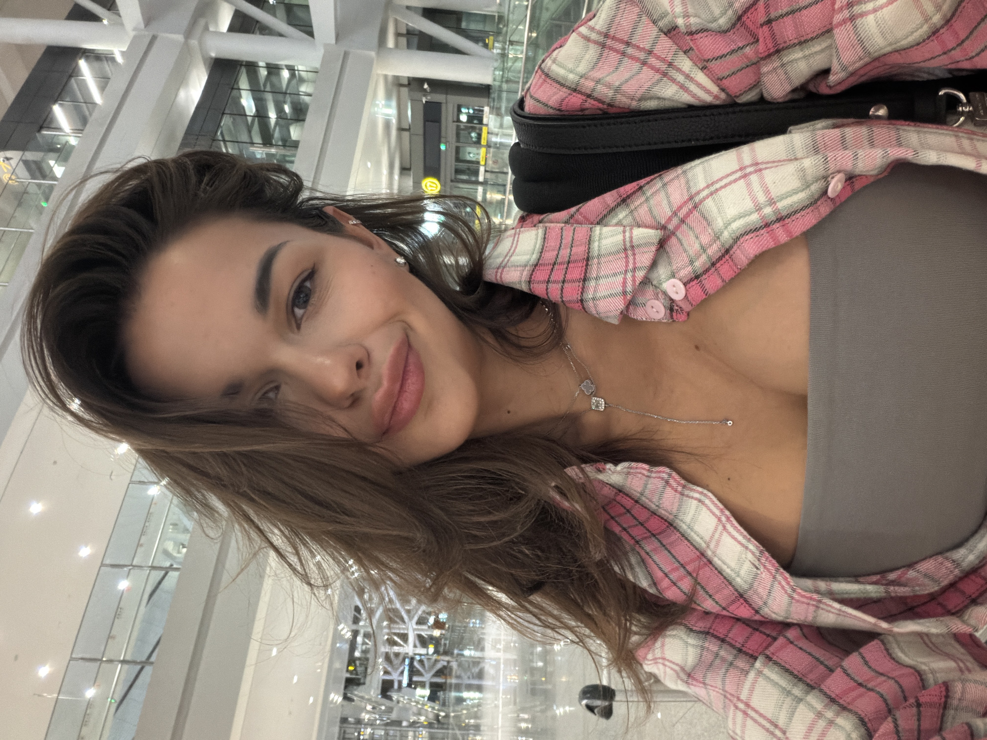 A woman takes a selfie in what appears to be an airport. She is wearing a gray top and a pink and white plaid shirt, along with a thin silver necklace. A black strap goes over her shoulder. The background shows airport architecture, including a glass wall, white beams, and indoor lighting. While it's not possible to confirm the exact location without further evidence, the architecture suggests a modern airport terminal. There is no verifiable information in the image to suggest the location is in Langfang, China.