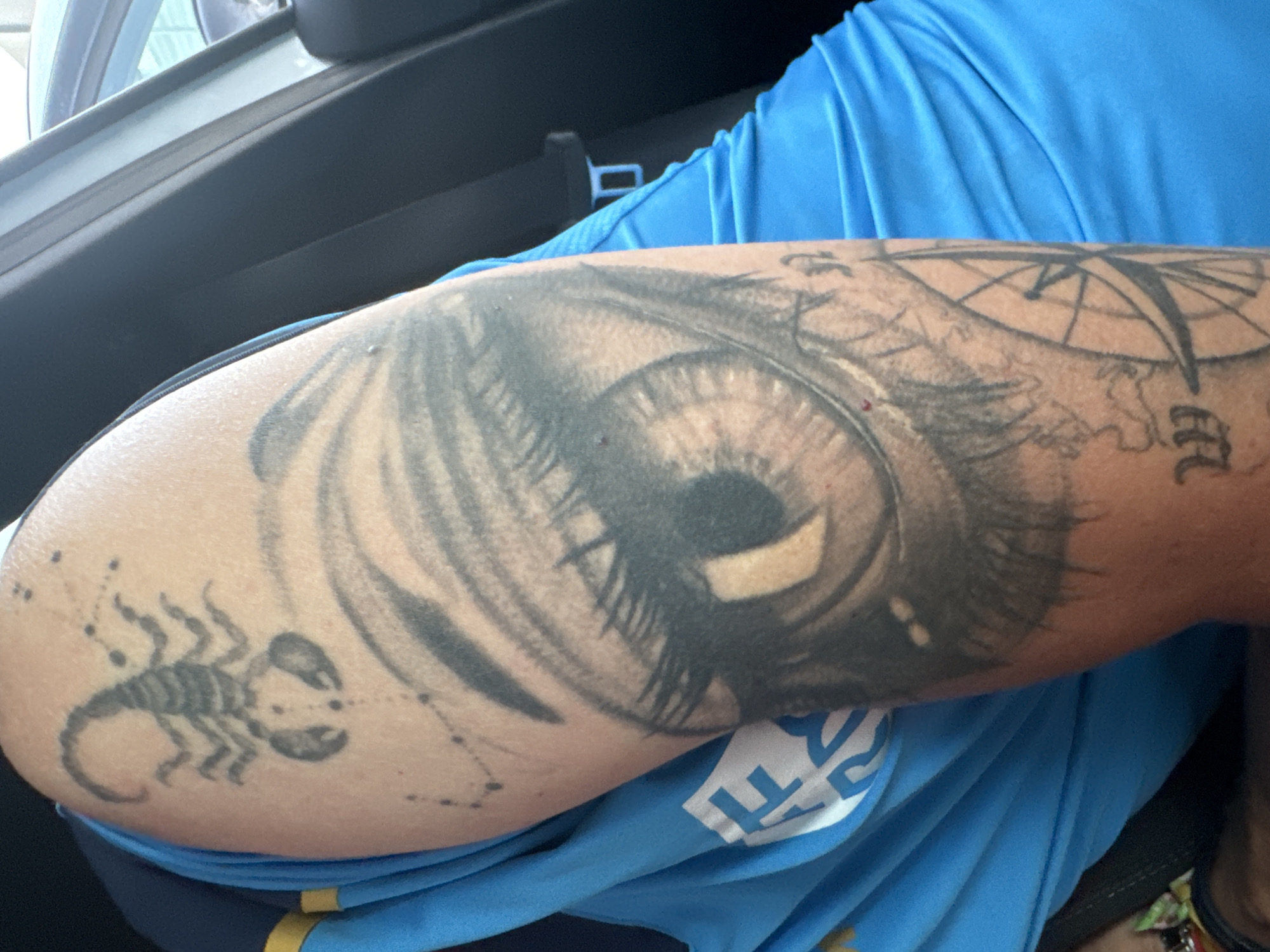 The image shows a close-up of a person's arm with tattoos, worn as they sit in the passenger seat of a vehicle. The tattoos are of a scorpion, a detailed eye, and what appears to be a compass or map. The person is wearing a bright blue shirt with yellow trim. The background shows the interior of a car and a blurred view of the outside. The specific location in the city of Murcia, Spain cannot be determined from the provided image.