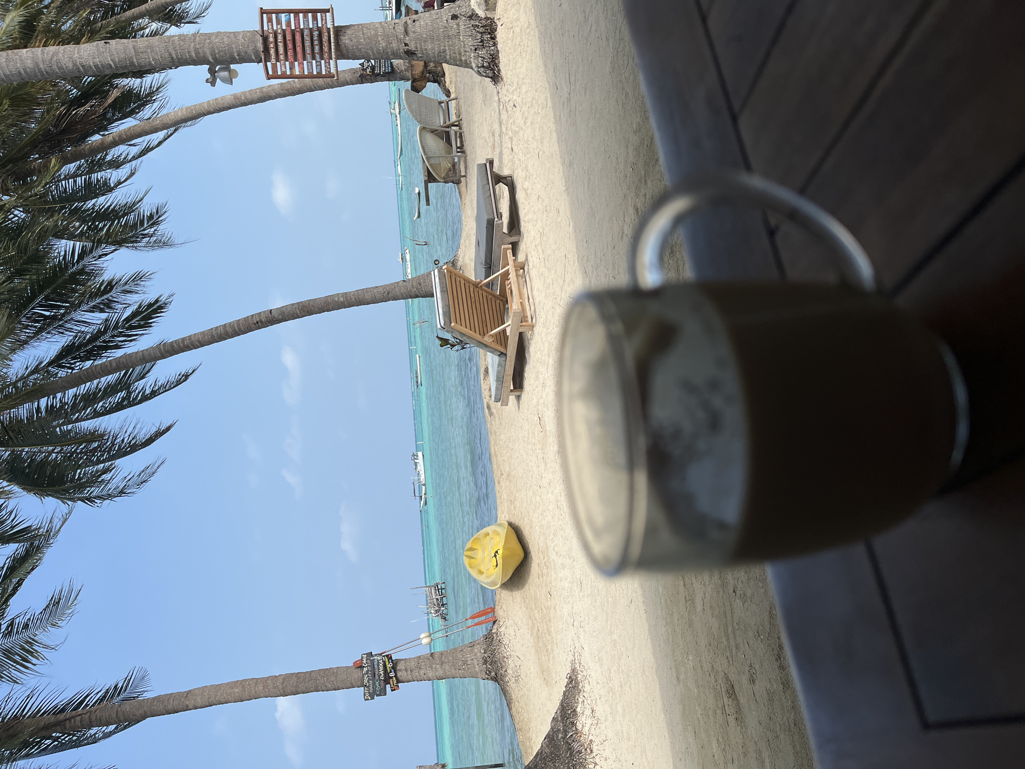 The image depicts a serene beach scene in Legonipah, Indonesia, with a clear blue sky overhead and lush palm trees framing the view. In the foreground, a blurred glass mug filled with a light brown beverage sits on a wooden surface. Beyond the mug, the sandy beach is visible, leading to the turquoise waters of the ocean. A bright yellow kayak rests on the sand near the water's edge. Several lounge chairs are arranged on the beach, inviting relaxation. In the distance, boats are anchored in the calm sea, and wooden structures suggestive of piers or small buildings are visible along the coastline. The overall atmosphere is peaceful and idyllic, typical of a tropical vacation destination.